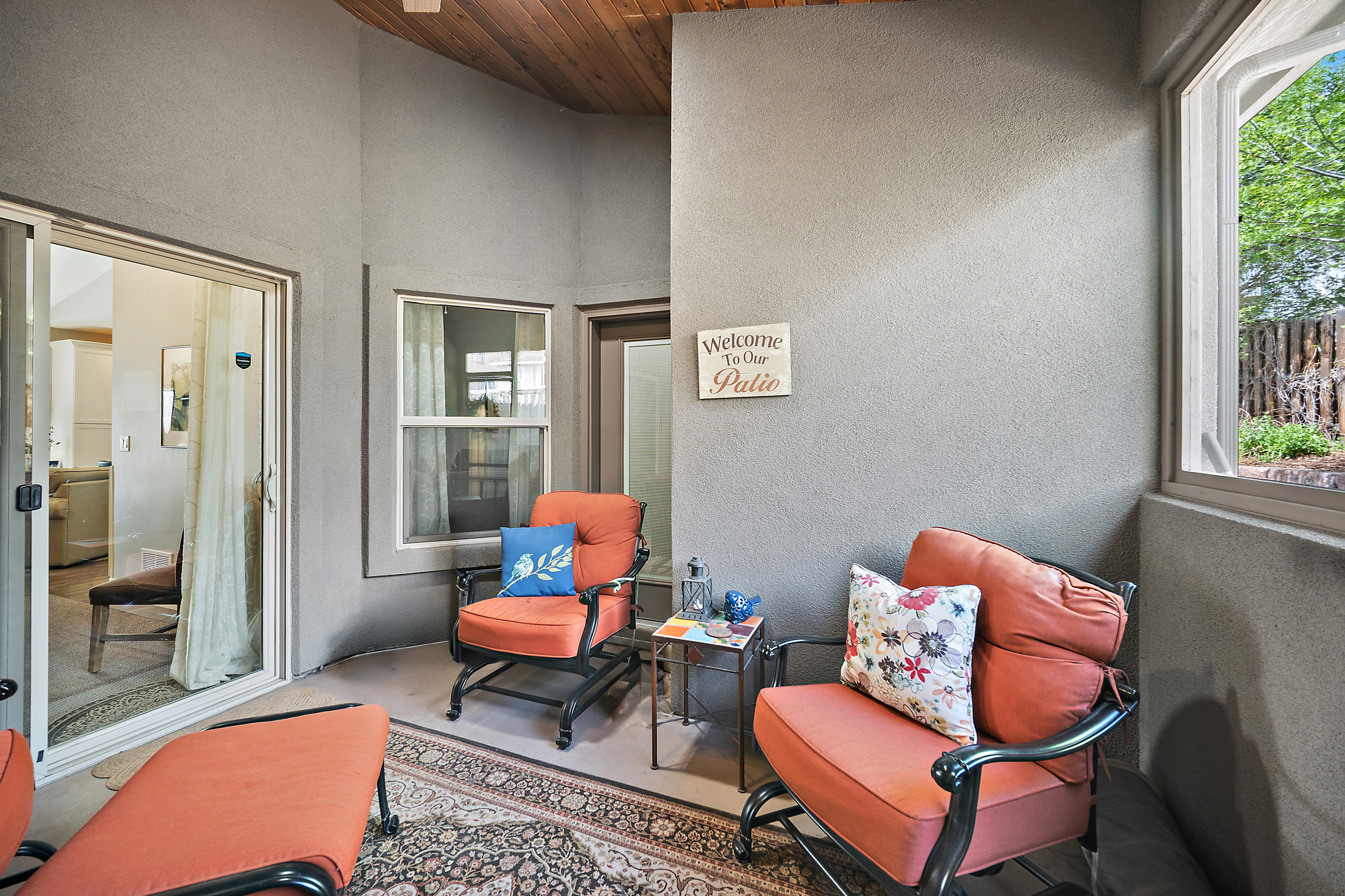 This cozy, covered patio space features a comfortable sitting area with orange cushioned chairs and matching footstools arranged on an ornamental rug. The space is highlighted by warm accent pillows, a decorative 'Welcome to Our Patio' sign, and a small side table, making it an inviting outdoor living area. A sliding glass door connects the patio to the interior, while a large window provides a view of the outside yard.