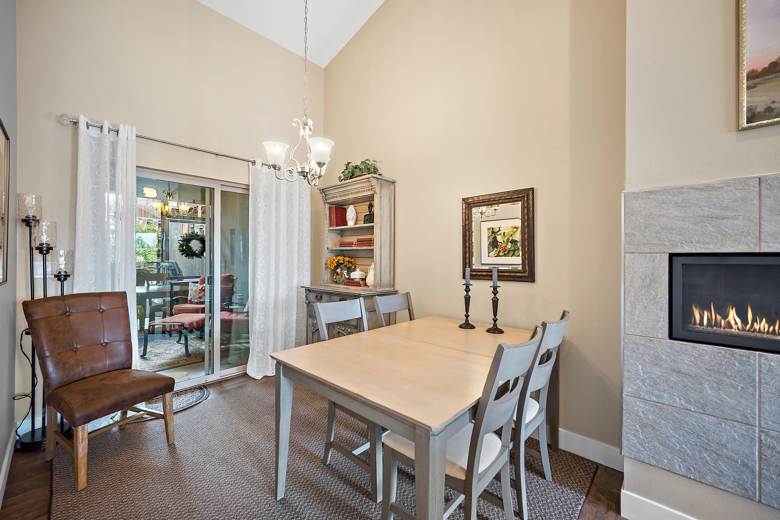 This bright and inviting dining area features a light-toned wooden dining table accompanied by gray chairs and an elegant light fixture overhead. A rustic hutch provides functional storage and adds classic charm, while the adjoining fireplace with stone tiling creates a cozy, focal point for the space. The room is completed by a sliding glass door that leads to an outdoor patio, enhancing the flow and natural light in the home.
