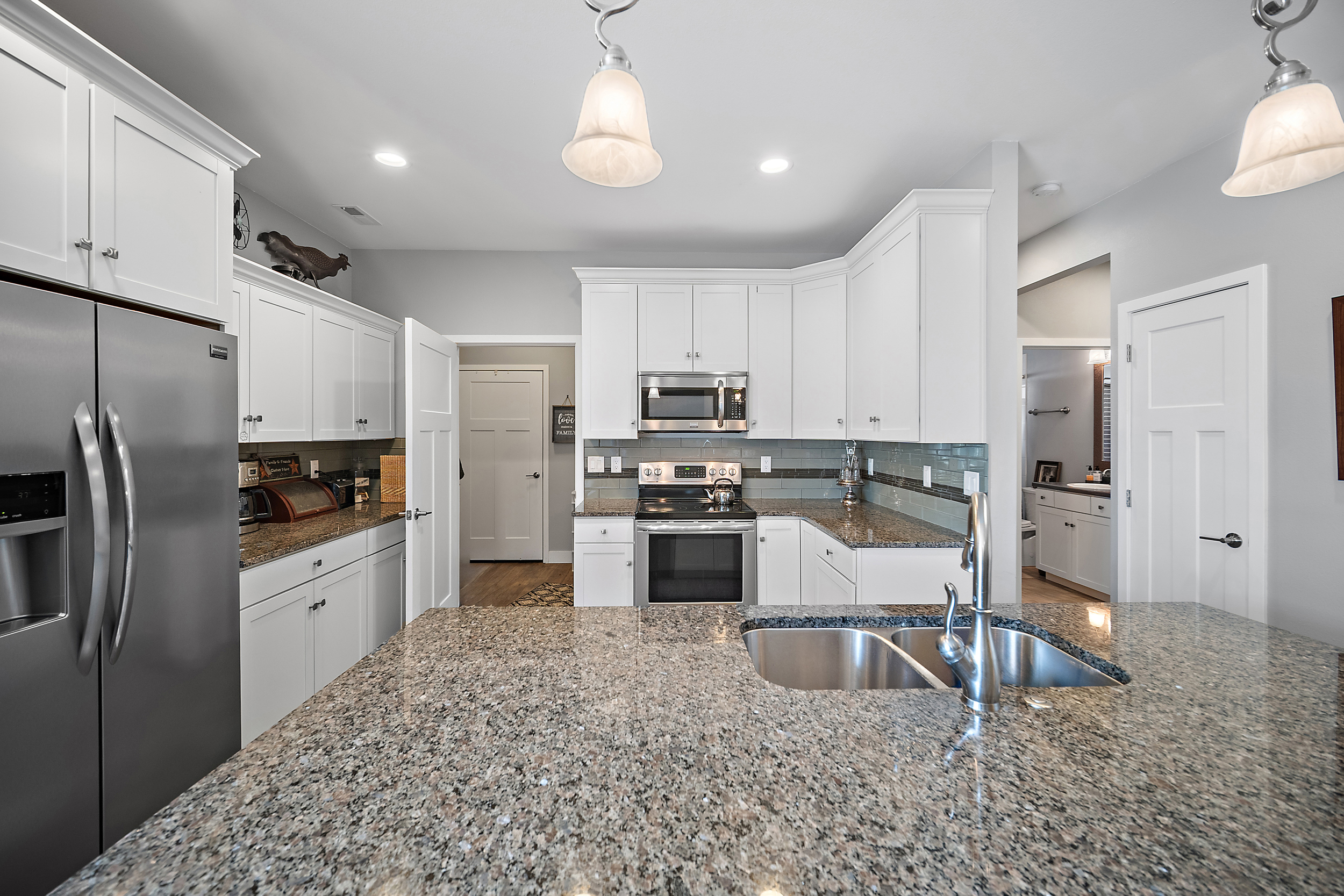 This bright, well-organized kitchen features clean white shaker-style cabinetry, granite countertops with a spacious center island, and stainless steel appliances. The layout includes a tiled backsplash and pendant lighting, providing a functional and modern aesthetic. The open design connects seamlessly to adjacent areas, highlighting a comfortable and inviting atmosphere ideal for both daily living and entertaining.