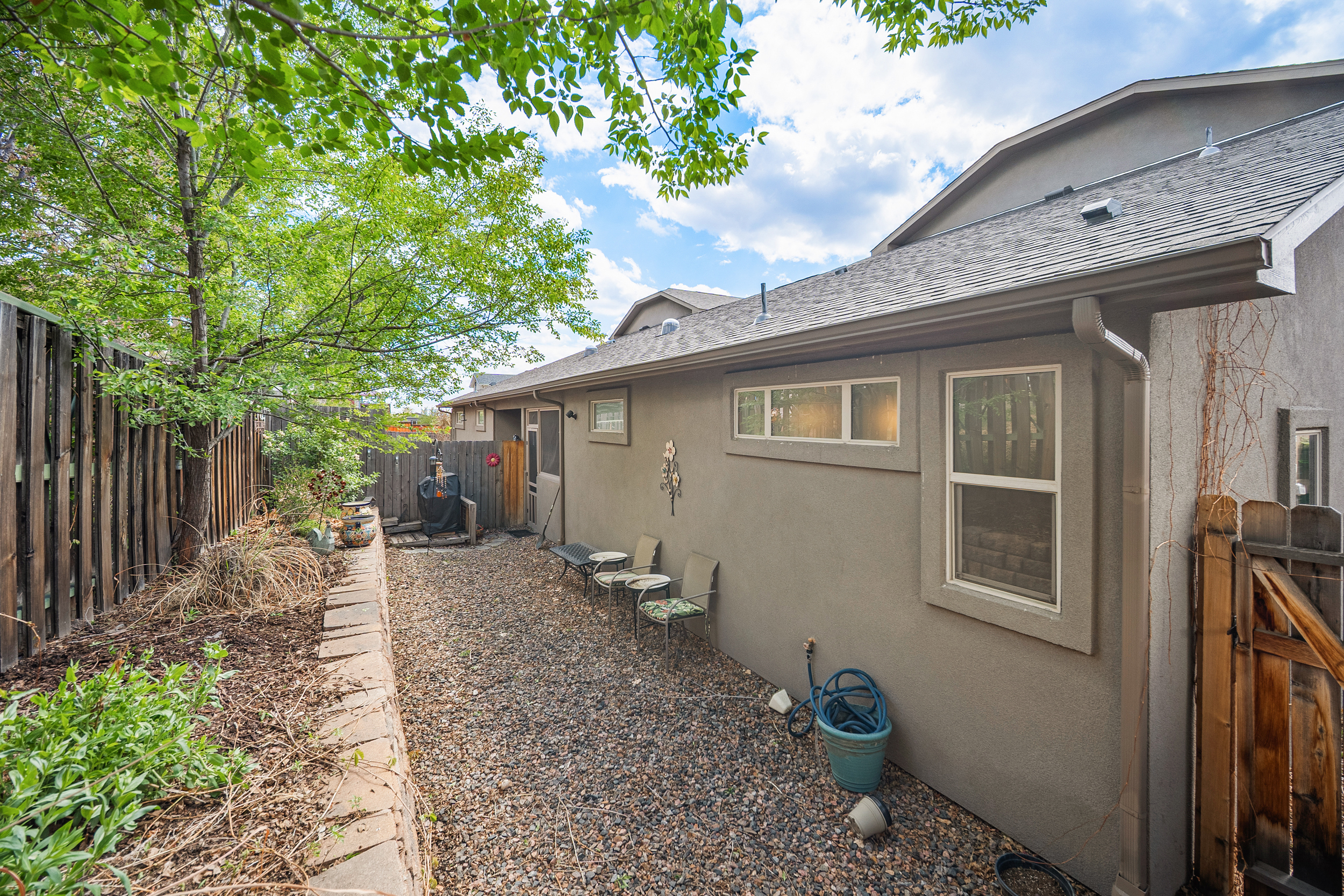 This image shows the side yard of a residence, featuring a stone-lined garden bed, a gravel pathway, and a stucco exterior wall. The area includes a small, casual seating arrangement and a hose stored in a bucket, suggesting a low-maintenance, functional outdoor space. A large tree provides shade, while the wooden fence and adjacent house structure frame the narrow side yard.