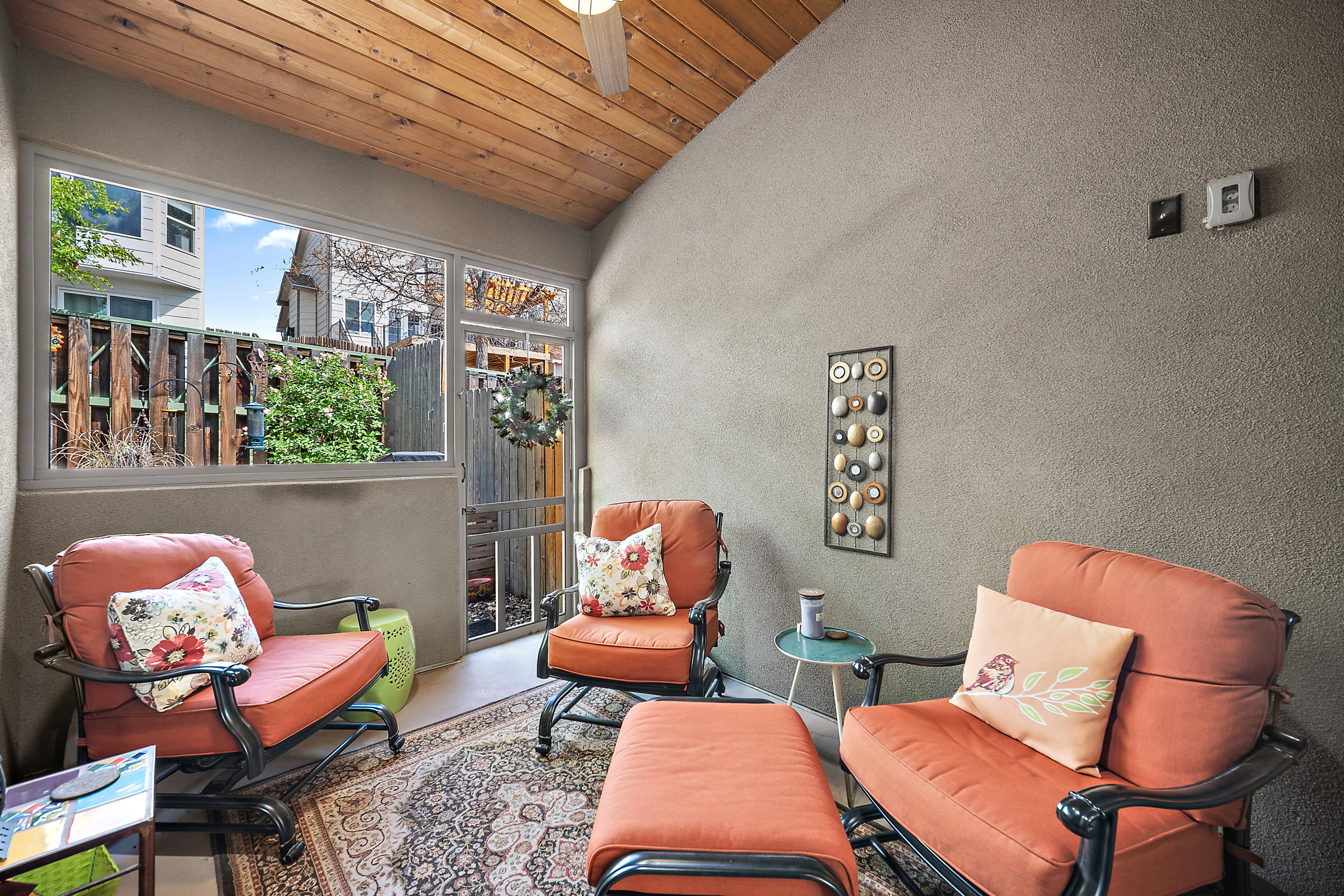 This cozy sunroom features a warm, wooden plank ceiling and textured walls, creating a welcoming and comfortable atmosphere. It is furnished with three bright orange patio-style chairs, a matching ottoman, and a patterned area rug, making it an ideal space for relaxing. A large window and a screen door provide views of the backyard, filling the room with natural light and connecting the indoor space with the outdoors.