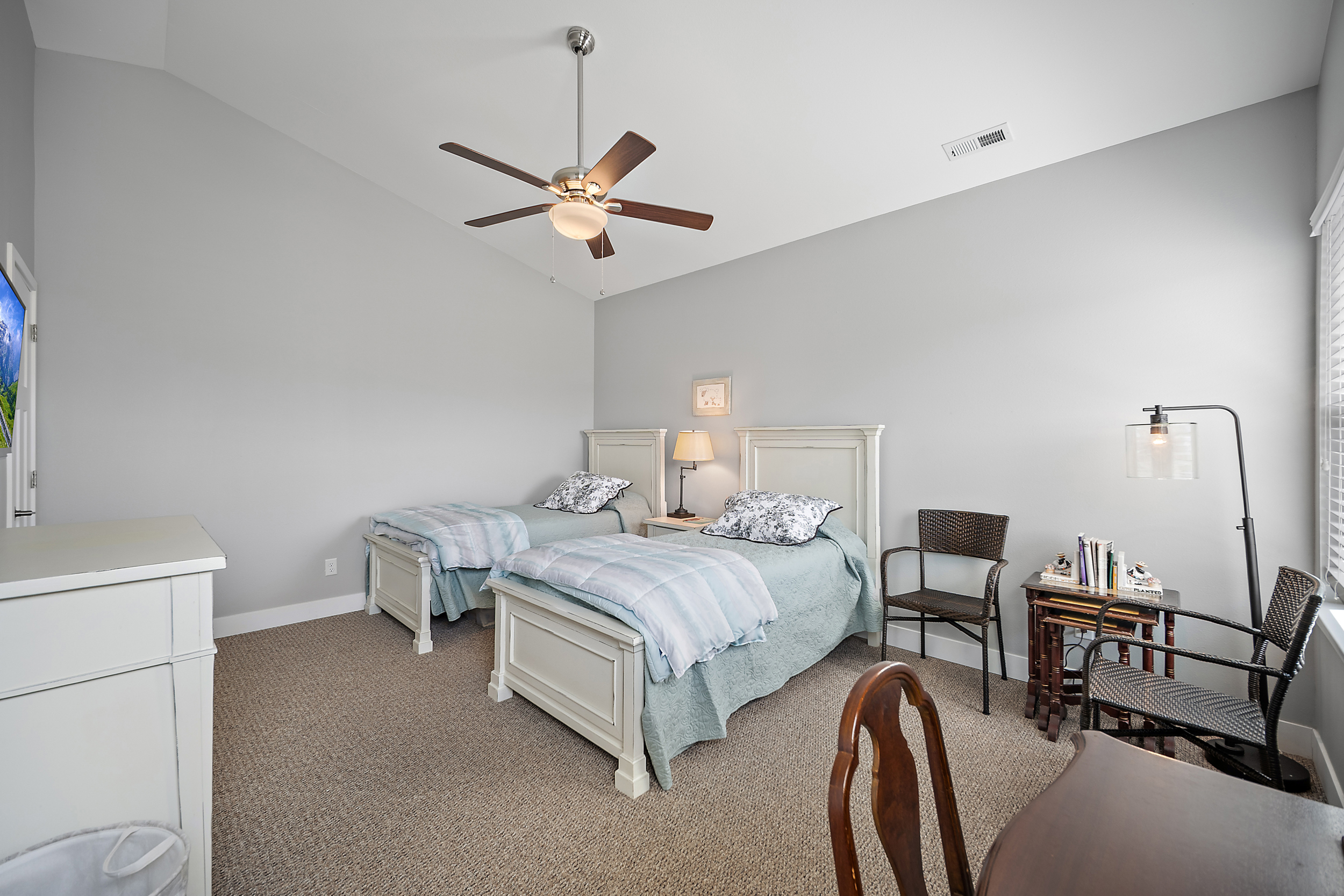 This guest bedroom features two matching white twin beds with complementary blue and white bedding, creating a bright and welcoming atmosphere. The neutral gray walls and carpeting provide a versatile backdrop, while an integrated ceiling fan and functional furniture, including a bedside table and seating area, add comfort and utility. Large windows allow for ample natural light, making the room feel spacious and well-lit.