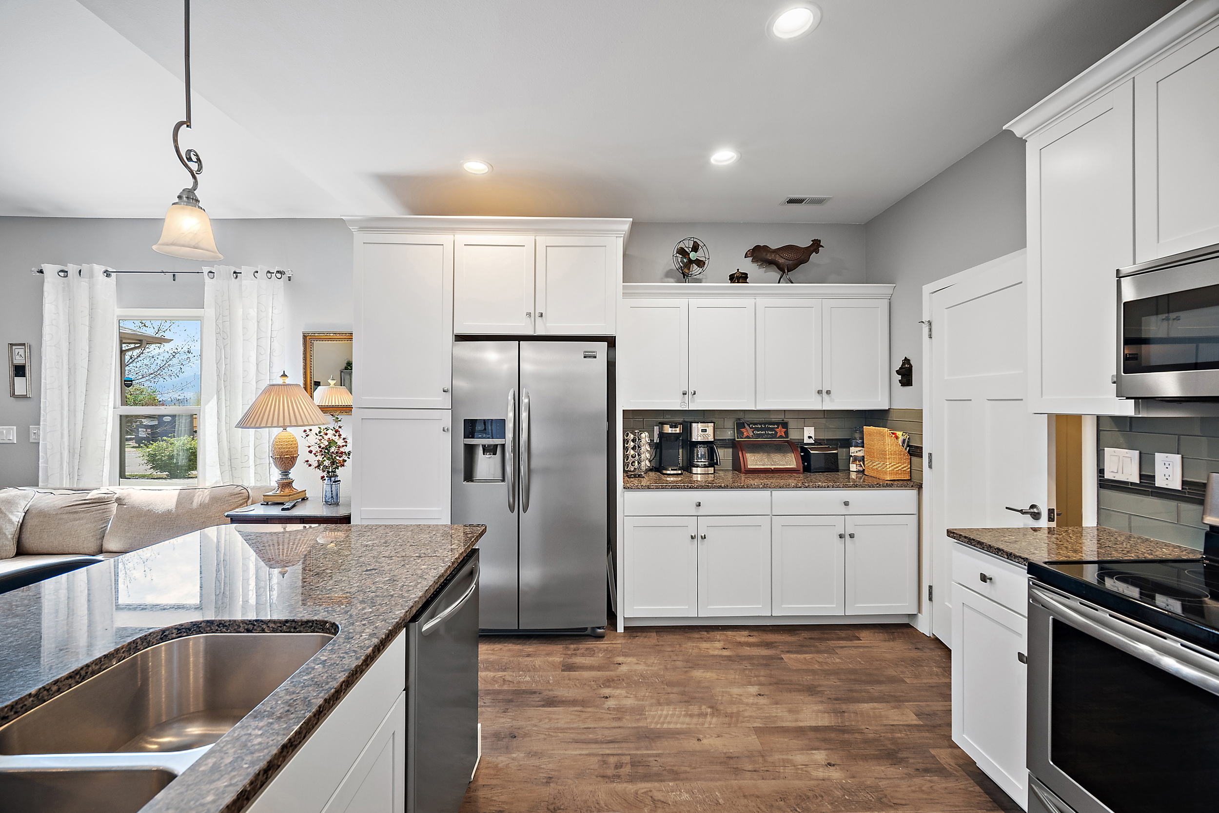 This modern, bright kitchen features clean white shaker-style cabinetry, high-end stainless steel appliances, and contrasting granite countertops. The space boasts a functional layout with a spacious island for seating, warm wood-toned flooring, and a subway tile backsplash. Stylish pendant lighting and ample natural light from the adjoining living area window create an inviting and well-appointed atmosphere.