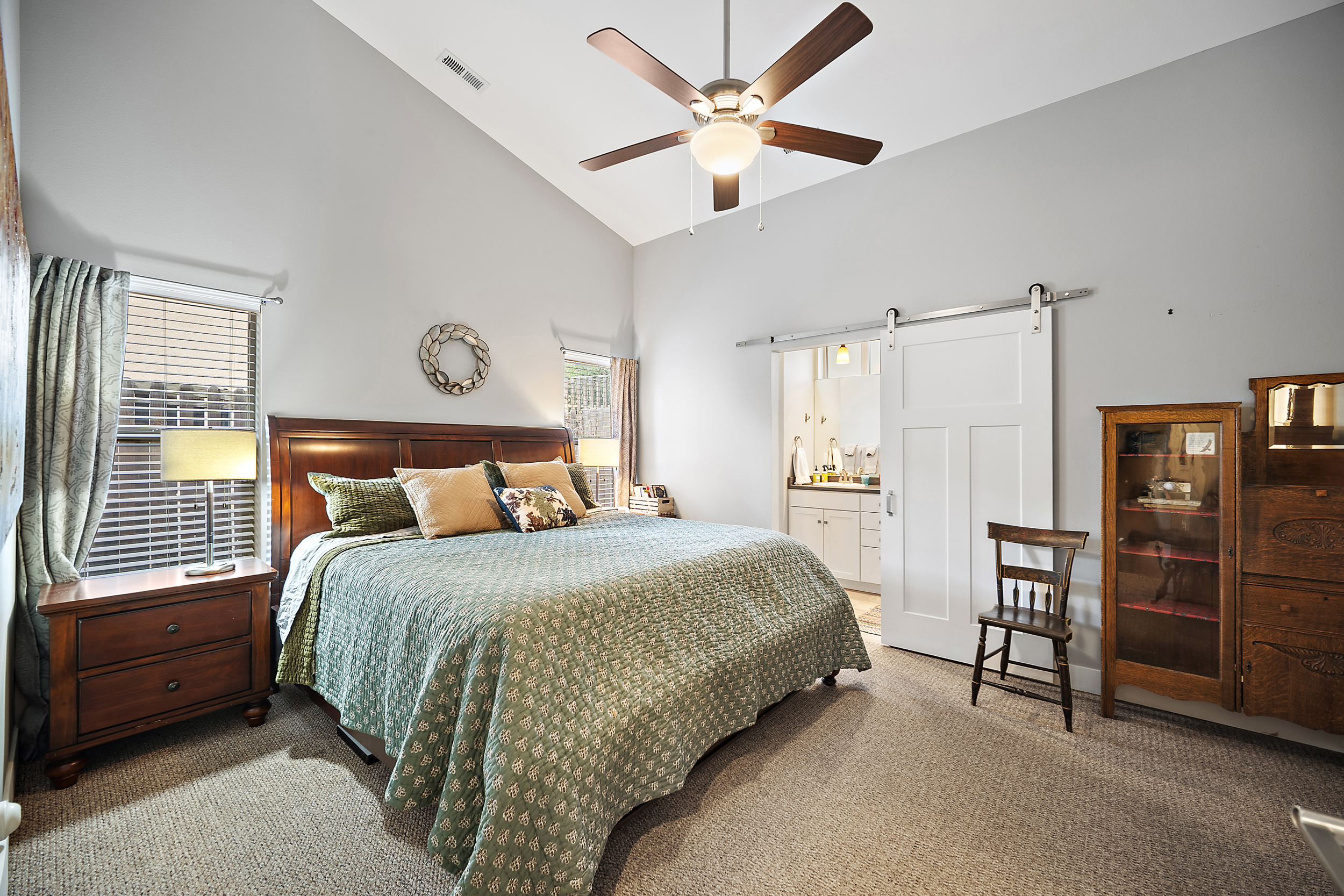 This spacious primary bedroom features a vaulted ceiling with a central ceiling fan and neutral wall tones. The room includes a large bed with ornate wooden headboard, a cozy seating area with a vintage-style wooden chair, and a sliding barn door that opens into an en-suite bathroom. A wooden curio cabinet and matching nightstand with a lamp enhance the room's traditional and welcoming atmosphere.