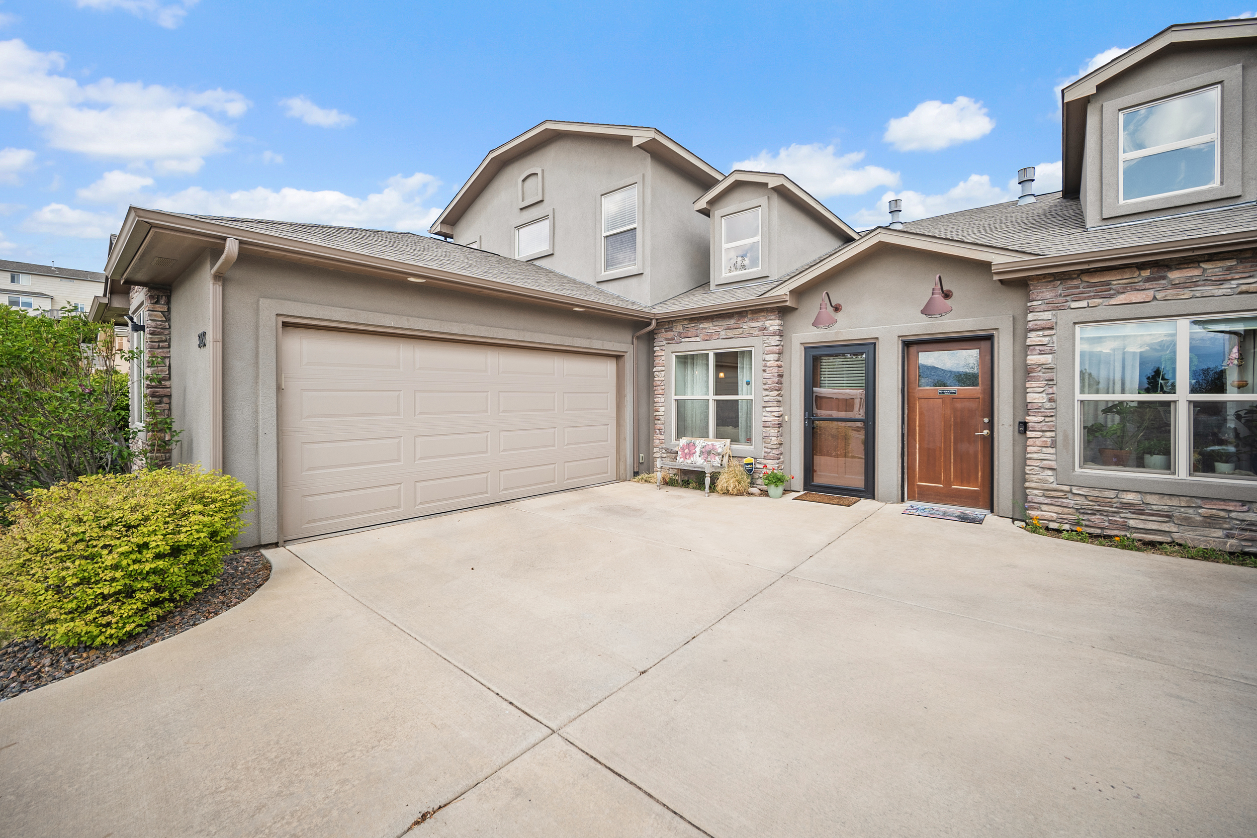 This attractive two-story residence features a charming combination of stucco siding and natural stone veneer accents. The front entrance is highlighted by dual wall-mounted light fixtures, while a spacious concrete driveway leads to an attached double-car garage. A well-maintained garden area sits to the left, contributing to the property's inviting and cohesive curb appeal.