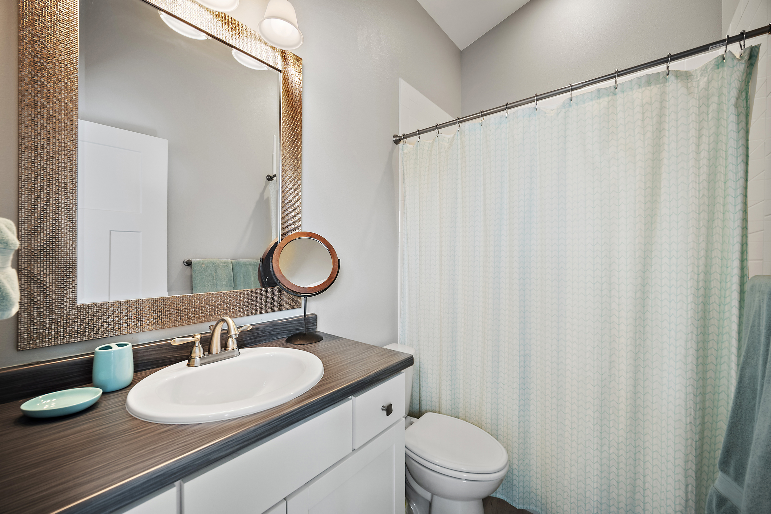 This clean and functional bathroom features a white vanity topped with a dark wood-look laminate countertop and a drop-in sink. A textured, metallic-style framed mirror hangs above the vanity, complemented by a light-colored chevron print shower curtain that adds a touch of modern personality. The space is well-lit and tidy, providing a bright and welcoming atmosphere.