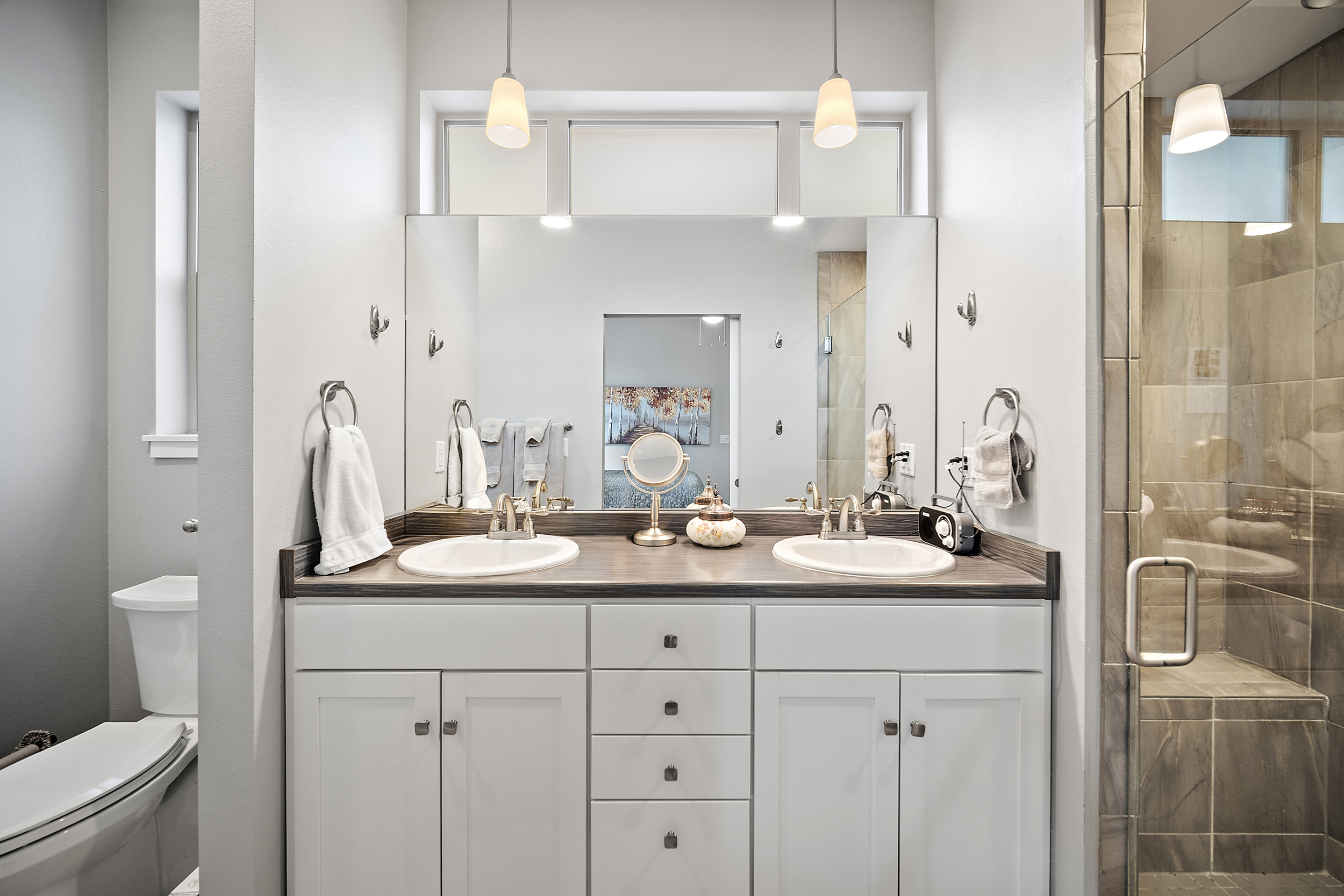 This primary bathroom features a double-vanity with white cabinetry, wood-look countertops, and dual undermount sinks. The space includes a large wall-to-wall mirror, elegant pendant lighting, and a walk-in tiled shower with glass doors. Neutral wall colors and classic hardware give the room a clean, functional, and well-maintained aesthetic.