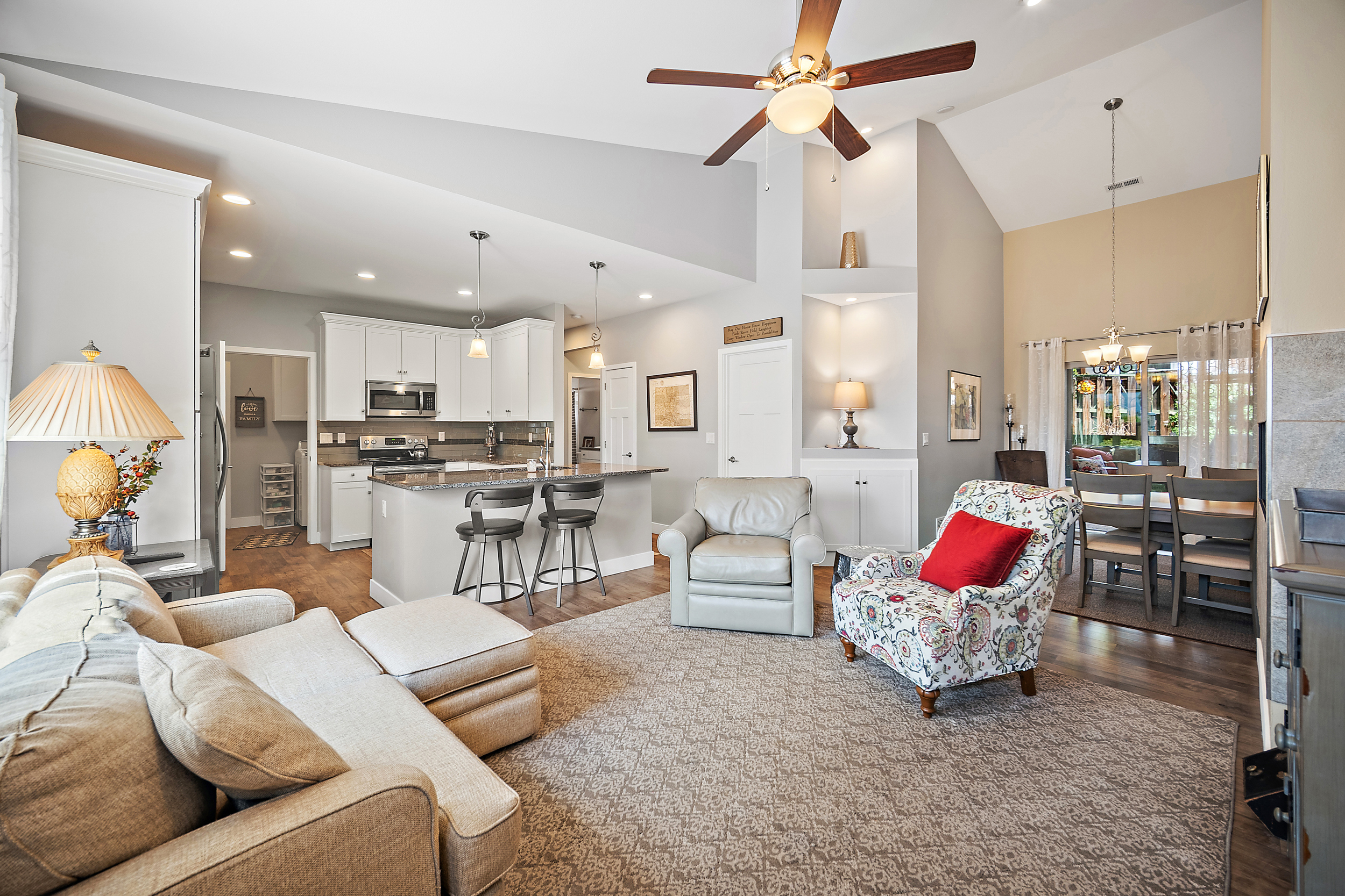 This open-concept living area features a vaulted ceiling with a fan, seamlessly blending into the kitchen and dining room. The space is neutrally styled with a comfortable sectional, a patterned accent chair with a red pillow, and a beige leather armchair atop a soft area rug. With its warm lighting, neutral color palette, and intuitive flow, the home presents a welcoming and cohesive living environment suitable for modern entertaining.