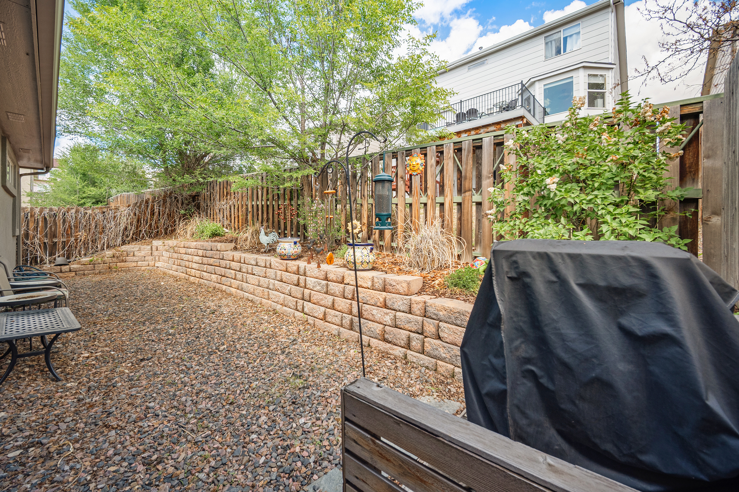 This backyard features a tiered retaining wall made of stacked stone blocks, creating a raised garden bed filled with shrubs and decorative items. The ground area is laid with gravel, providing a low-maintenance patio space adjacent to a covered outdoor seating area and a grill. A wooden fence provides privacy, while a mature tree adds shade and natural appeal to this functional outdoor living space.