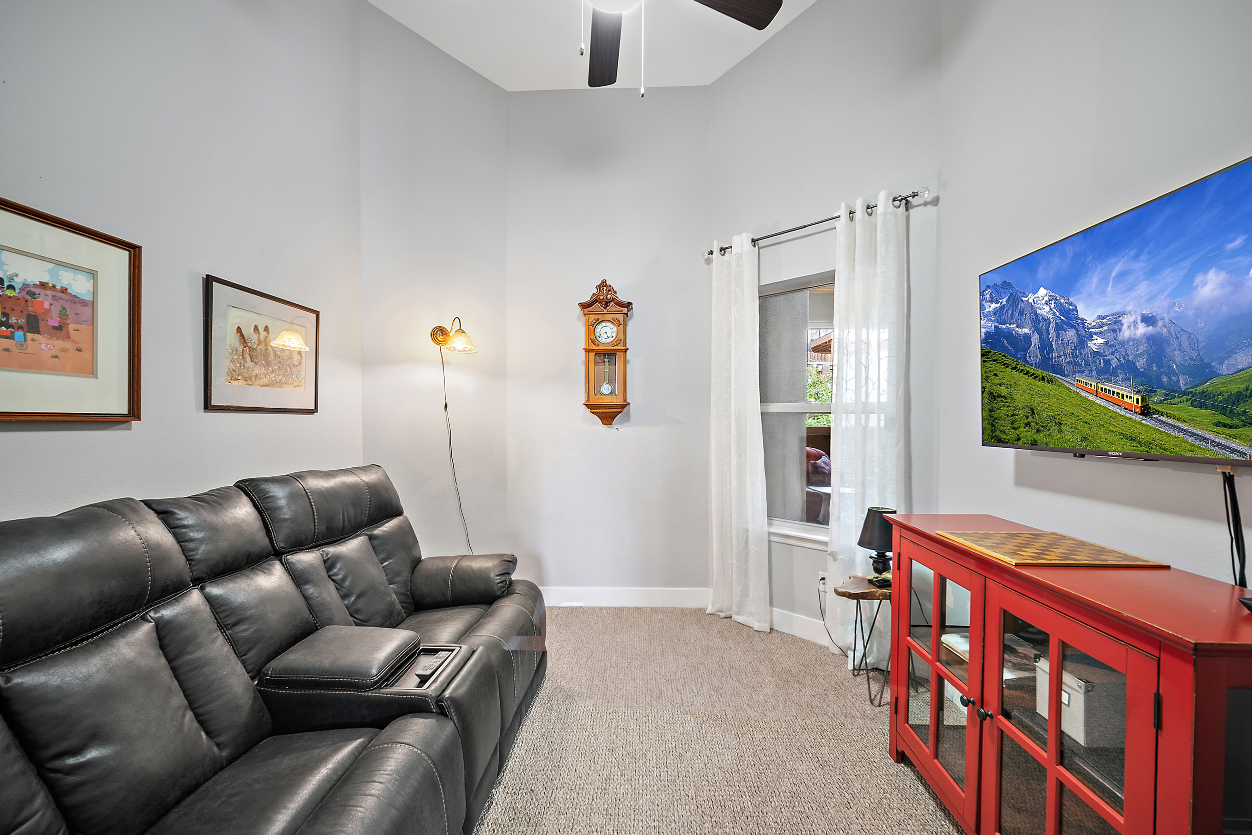 This cozy living area features a dark leather reclining sofa paired with a vibrant red media console, creating a comfortable space for relaxation. The room is accented by a classic wall-mounted grandfather clock, a stylish wall sconce, and framed artwork, all topped by a soaring vaulted ceiling. A large flat-screen TV provides entertainment, while a window with sheer curtains allows natural light to brighten the space.
