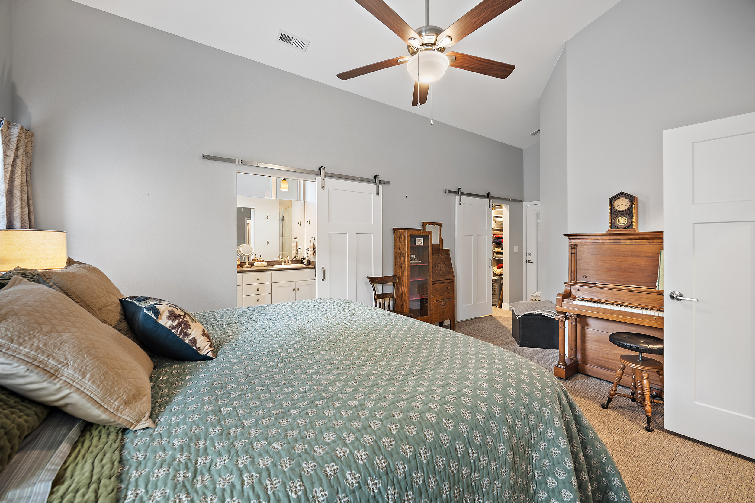 This spacious primary bedroom features a large, patterned quilt on the bed and an open-concept layout that flows into an en-suite bathroom and a walk-in closet through sliding barn doors. A wooden piano with a matching stool sits gracefully in the corner, adding a unique, traditional touch to the room. The space is topped off with a high vaulted ceiling and a ceiling fan, creating a welcoming and comfortable atmosphere.