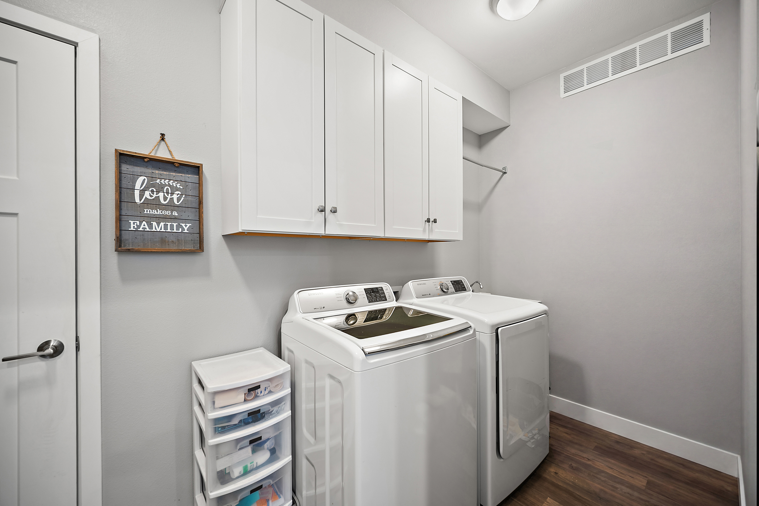 This laundry room features white upper cabinetry providing organized storage, paired with a modern top-loading Samsung washer and a matching dryer. The room has neutral grey walls, white baseboard trim, and dark wood-look plank flooring, creating a clean and functional space. A small decorative sign and a compact plastic storage unit add a touch of personality and utility.
