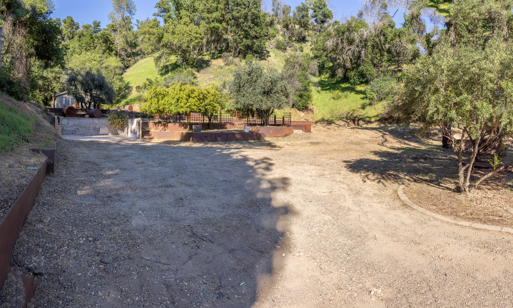 This outdoor space features a level dirt clearing that leads into a landscaped terrace with trees and a retaining wall. The area is bordered by mature trees and natural hillside vegetation, suggesting a rural or semi-rural property with significant potential for gardening or outdoor activities. The presence of metal retaining walls and deliberate landscaping steps indicates a clear structural design for this elevated section of the yard.