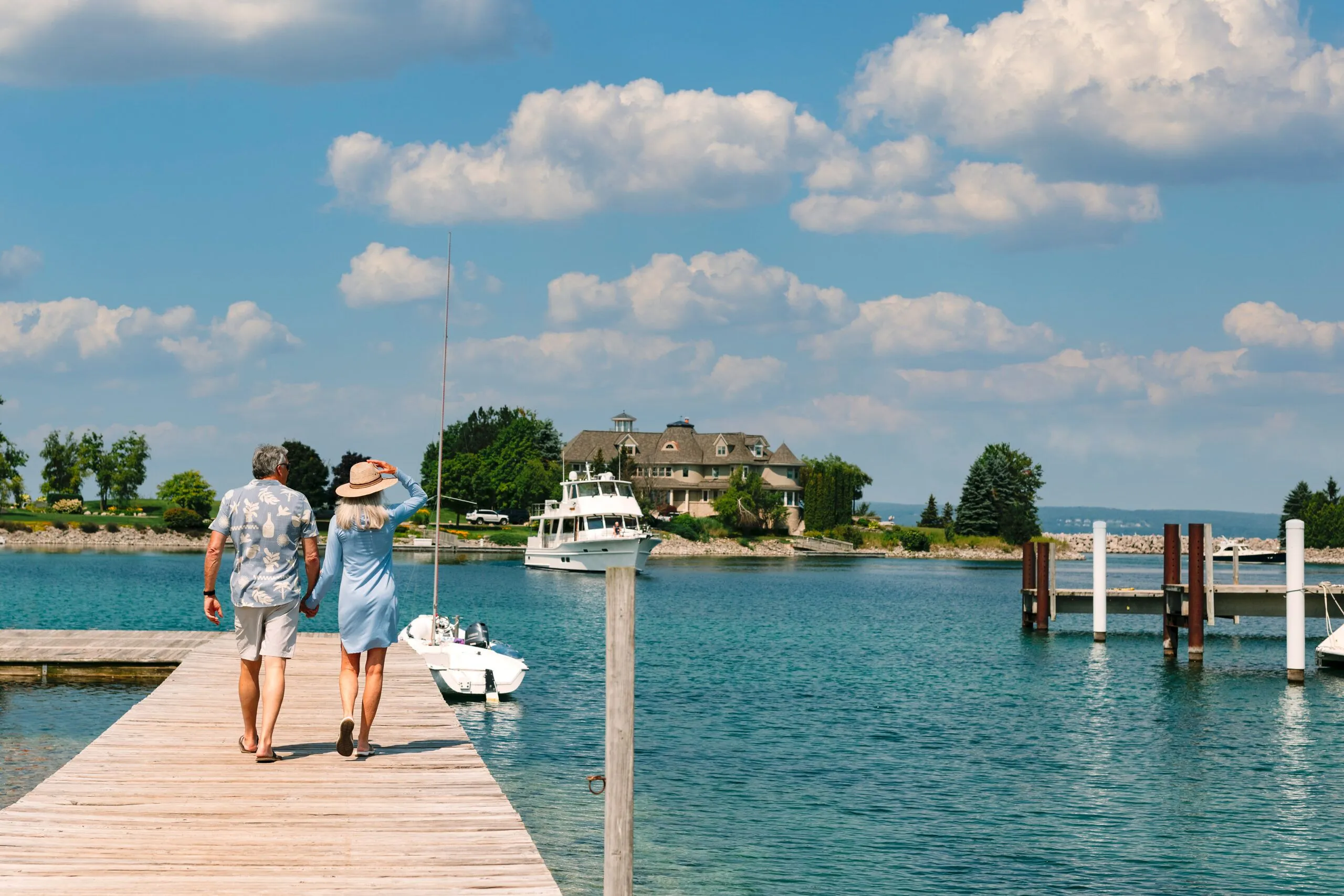 This image captures a picturesque waterfront setting featuring a long wooden dock leading toward a large, elegant estate home. The scene highlights access to boating and lakefront living, complete with a clear blue sky, a moored cabin cruiser, and a serene, upscale environment. It perfectly showcases the leisure-oriented lifestyle and natural beauty associated with high-end waterfront residential communities.