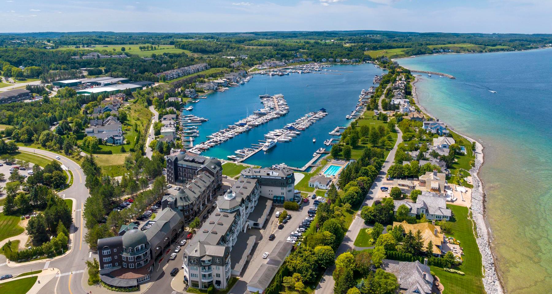 This stunning aerial view captures a luxury resort and marina complex nestled along a scenic shoreline. The property features expansive architectural buildings, a pristine swimming pool, and a well-maintained marina filled with yachts, all surrounded by lush green landscapes. Its premier waterfront location and comprehensive resort amenities make it a highly desirable residential and vacation destination.