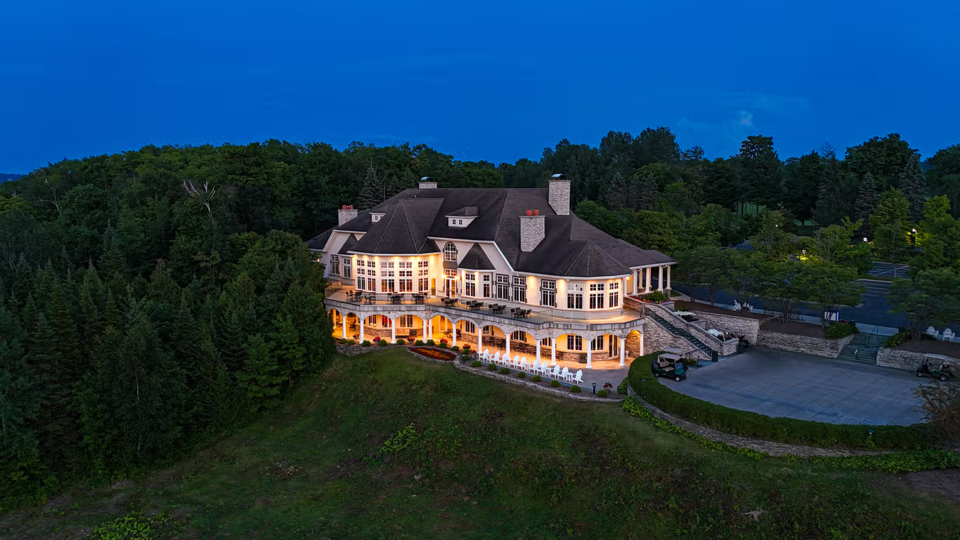 This is an impressive aerial view of a large, multi-story luxury estate nestled against a lush forest at twilight. The home features a grand, elevated terrace with decorative columns, warm lighting that accentuates its architectural details, and a sprawling paved driveway below. Its sophisticated design, expansive windows, and picturesque setting suggest a premium property, likely a clubhouse or exclusive residence.