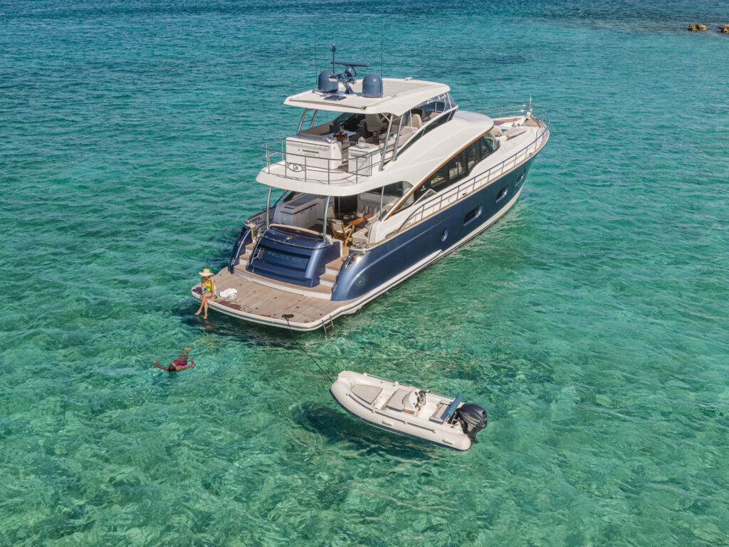 This high-angle, aerial shot captures a luxurious Belize 66 yacht anchored in crystal-clear turquoise waters. The vessel features a sleek dark blue hull, a spacious teak deck with an integrated swim platform, and a multi-level layout that includes an open-air upper deck. A small tender boat is tethered to the stern, and a person is visible swimming nearby, creating an inviting and idyllic scene suitable for luxury maritime lifestyle marketing.