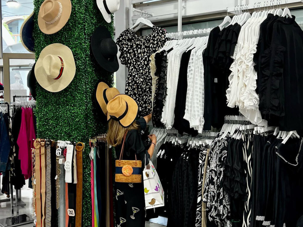 This image shows the interior of a boutique retail store featuring rows of clothing racks, organized displays of straw hats on a green faux-plant wall, and a selection of belts. A woman is seen browsing the racks, contributing to a vibrant and commercial atmosphere. The space is well-lit and neatly arranged, showcasing a variety of apparel and accessories.