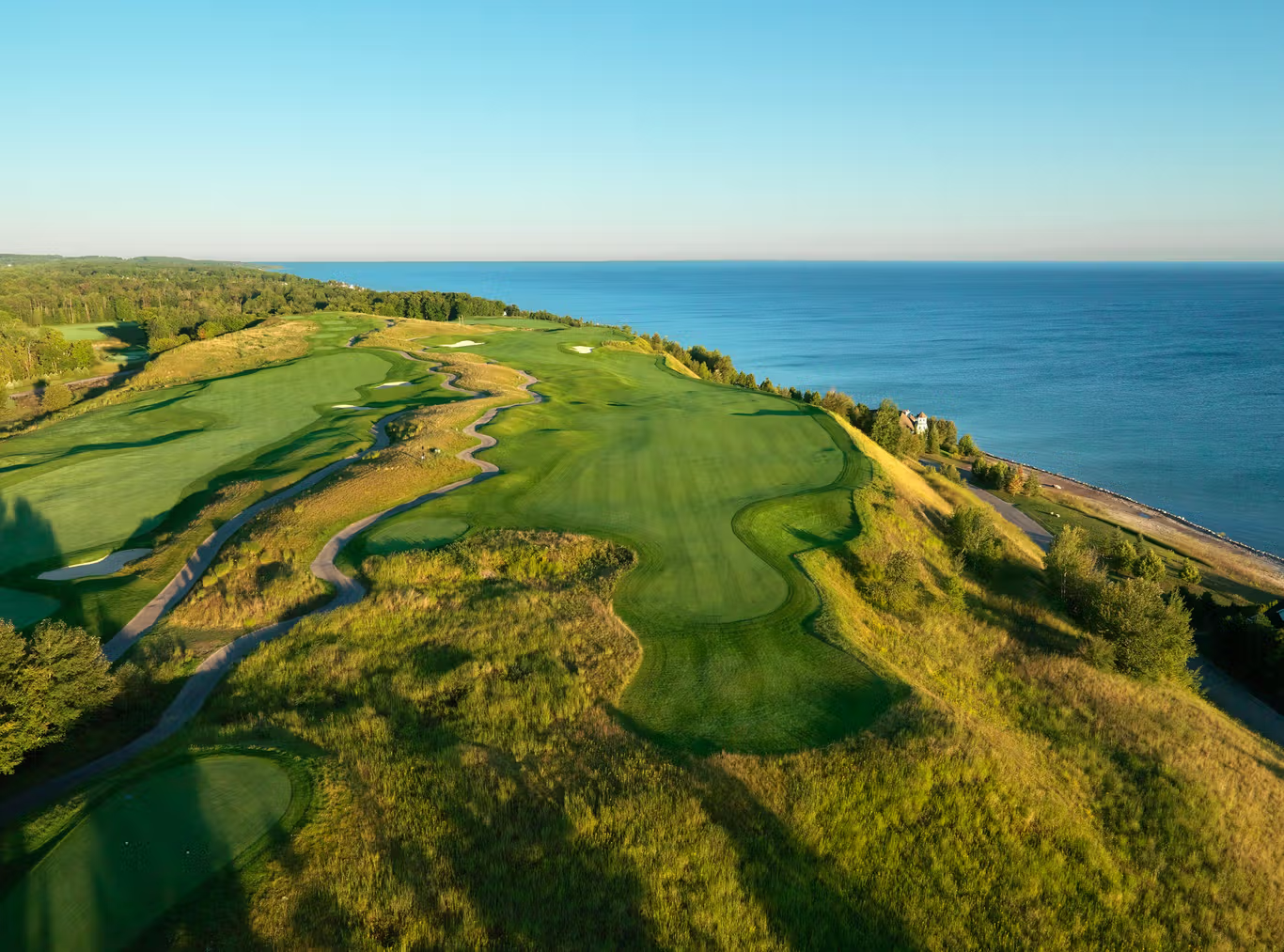 This stunning aerial view captures a premier golf course community located along a scenic coastline. The image highlights undulating fairways, meticulously maintained greens, and expansive grassy terrain that integrates seamlessly with the natural landscape and open water views. The property offers an exclusive lifestyle, blending leisure and outdoor recreation in a breathtaking, elevated setting.