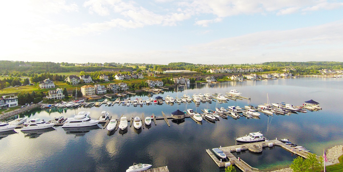This stunning aerial view captures a vibrant waterfront community featuring a sprawling marina filled with numerous docked yachts and boats. The landscape is dotted with picturesque multi-story homes nestled along the shoreline, creating a serene and upscale living environment. The clear, reflective water and well-maintained docks highlight the community's appeal as a premier destination for boating enthusiasts.