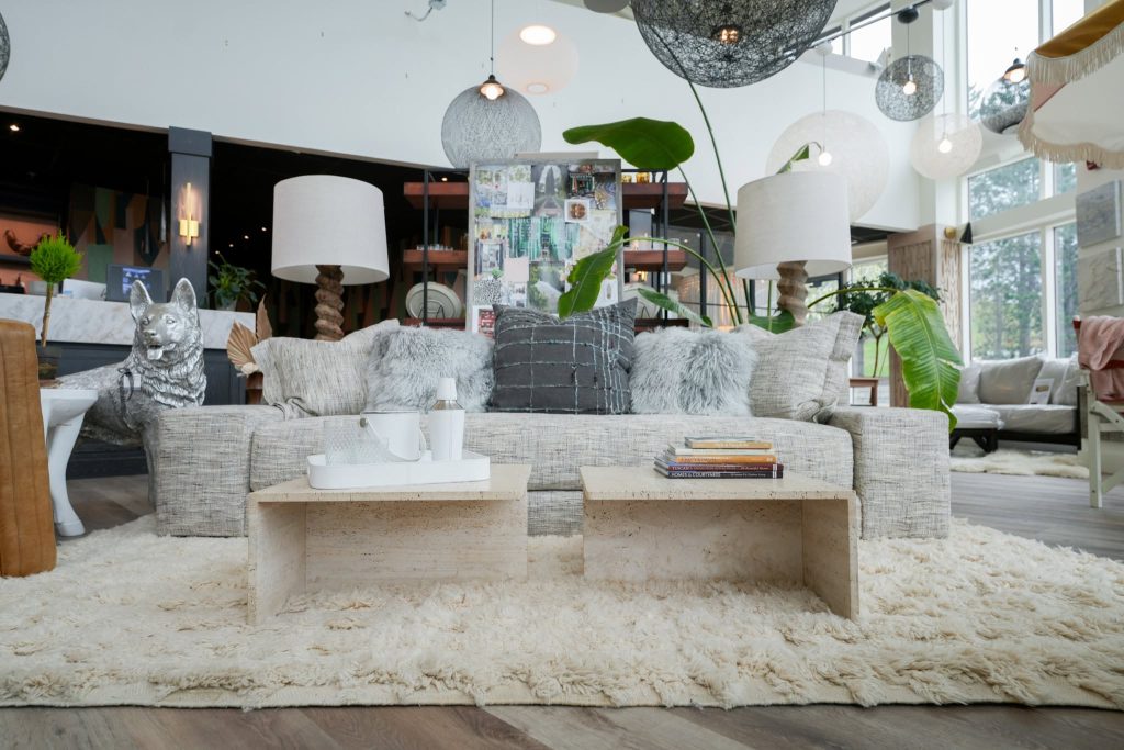 This living room area features a sophisticated, neutral-toned design centered around a large, textured sofa layered with plush pillows. Two minimalist travertine coffee tables ground the seating area, which is anchored by a high-pile cream area rug and illuminated by several unique, modern pendant lights. The space incorporates natural elements like indoor greenery and a decorative dog statue, creating a trendy, upscale, and inviting aesthetic.