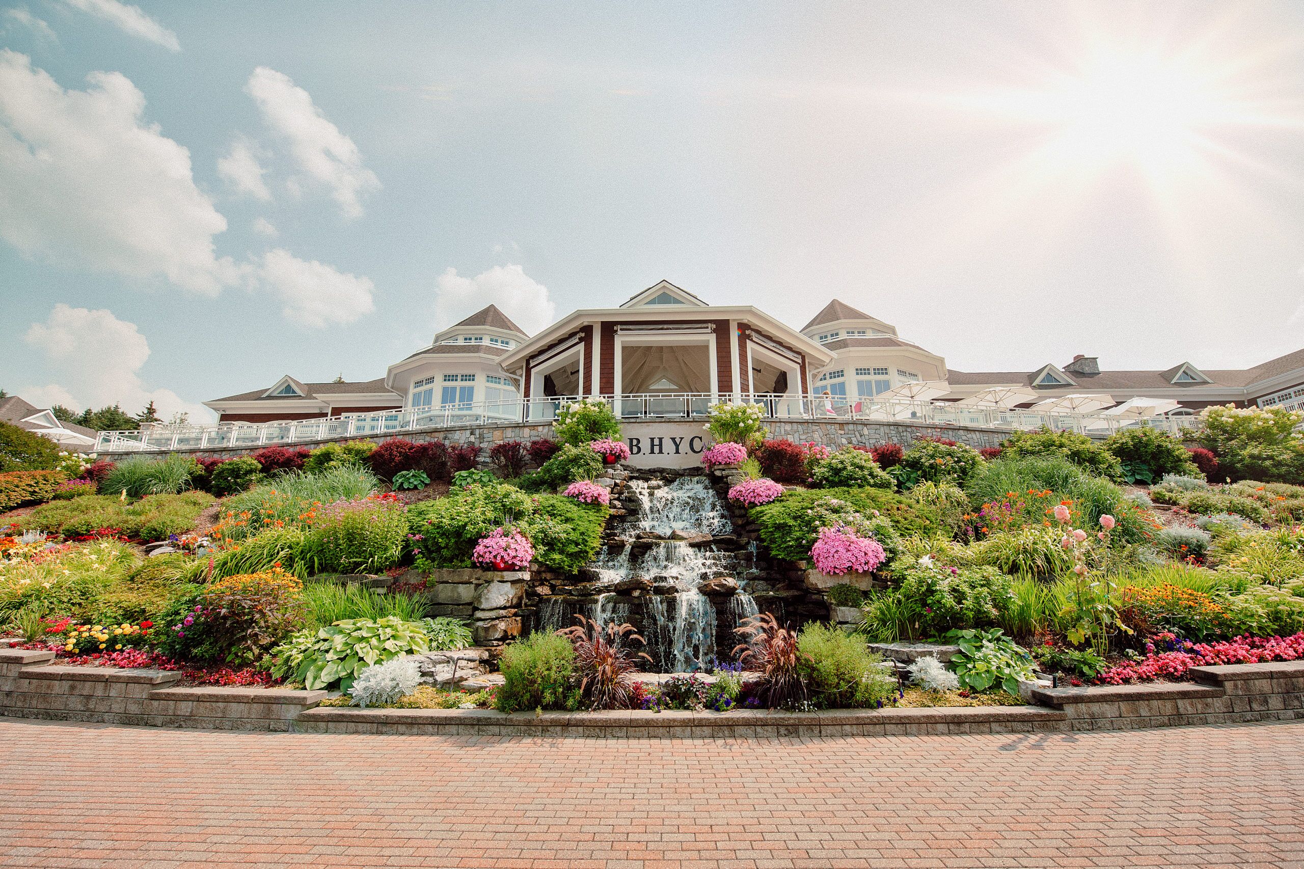 This image features the prominent exterior front facade of the B.H.Y.C. clubhouse, distinguished by its tiered stone retaining wall and an elaborate, multi-level water feature surrounded by lush, colorful landscaping. The architectural style includes prominent gables and expansive terrace railings, creating an inviting and well-manicured arrival experience. Its picturesque setting, highlighted by vibrant flower beds and a brick-paved foreground, conveys a sense of high-end community amenities.