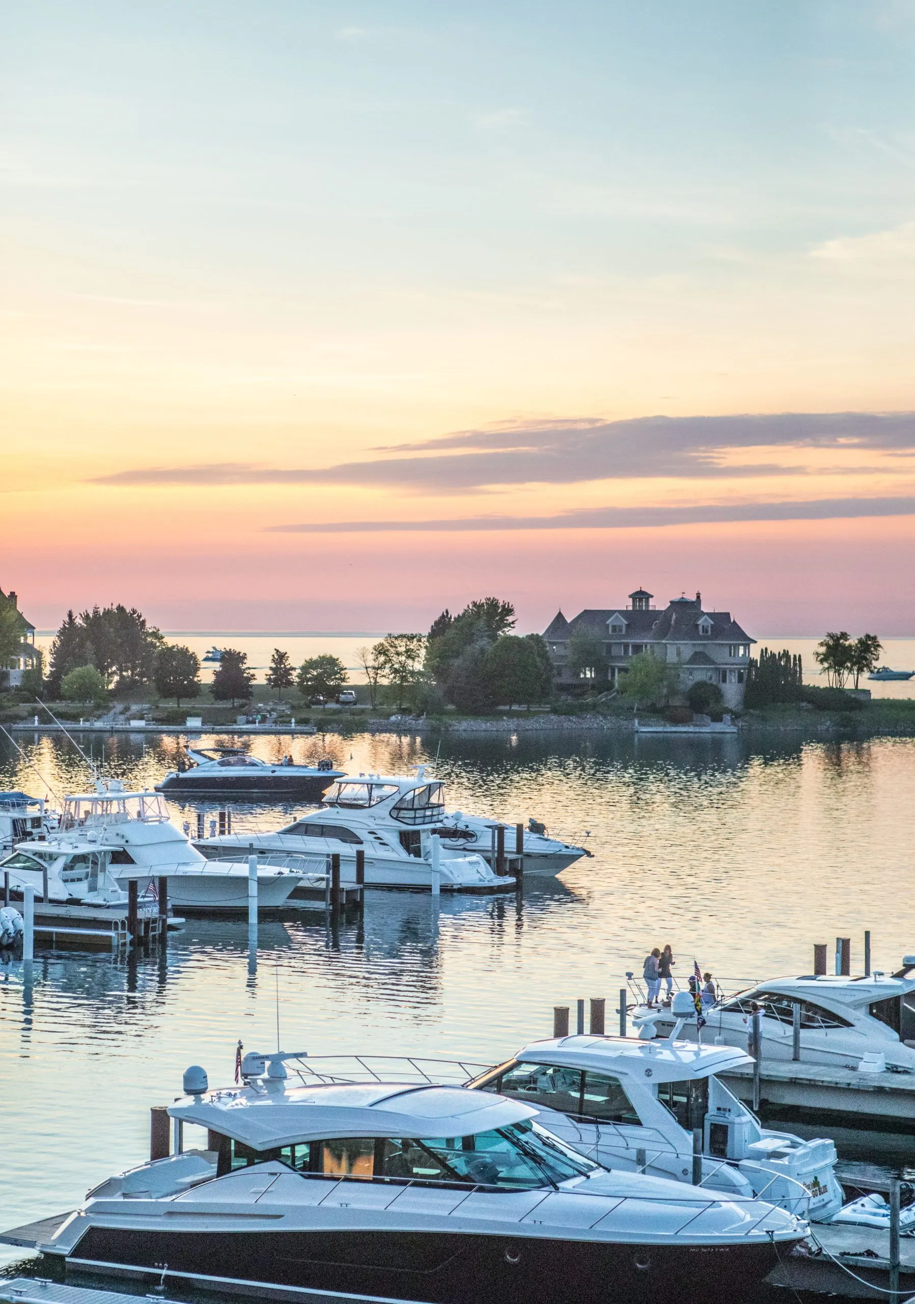 This scenic image features a private marina at sunset, showcasing high-end yachts moored at professional docks. In the background, a large, stately waterfront residence stands as a focal point, emphasizing the exclusive and luxurious lifestyle of this coastal community. The calm water, soft evening lighting, and proximity to luxury boats highlight the premium recreational amenities associated with this high-end residential setting.