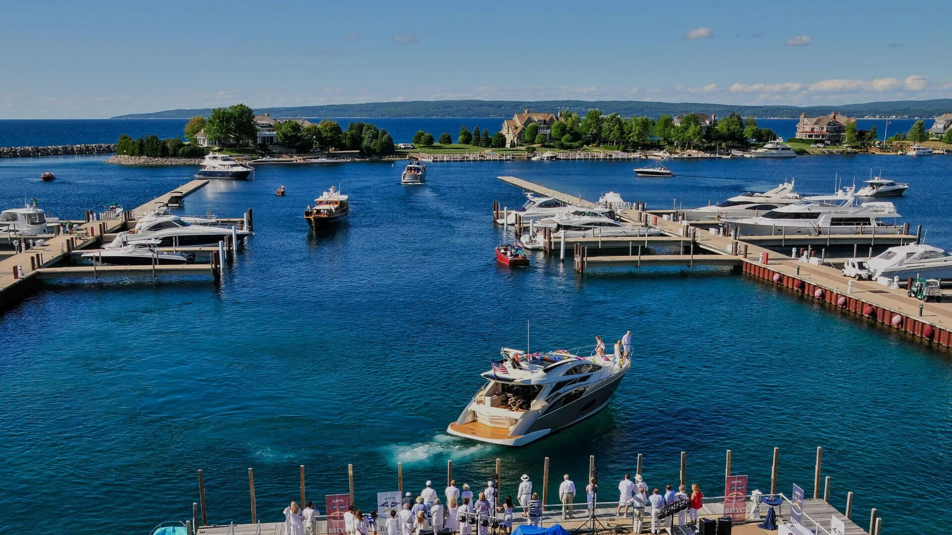 This aerial view showcases a vibrant harbor community featuring multiple docks filled with luxury yachts and boats. In the foreground, people gather on a pier to welcome an arriving yacht, highlighting the marina's active social scene and waterfront lifestyle. The surrounding landscape includes manicured greenery and upscale residences, reflecting an exclusive and desirable coastal neighborhood.