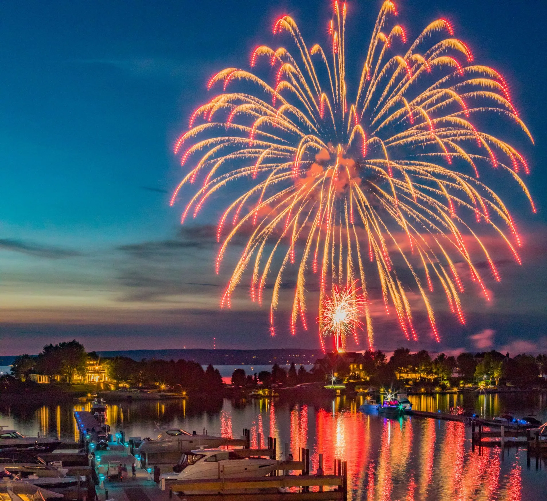 This stunning nighttime aerial view captures a vibrant waterfront scene illuminated by a spectacular fireworks display. The image features a busy private marina filled with docked boats, with luxury homes nestled along the shoreline reflecting colorful lights into the calm water. The atmospheric lighting and expansive perspective highlight the prime lifestyle and recreational appeal of this upscale lakeside community.