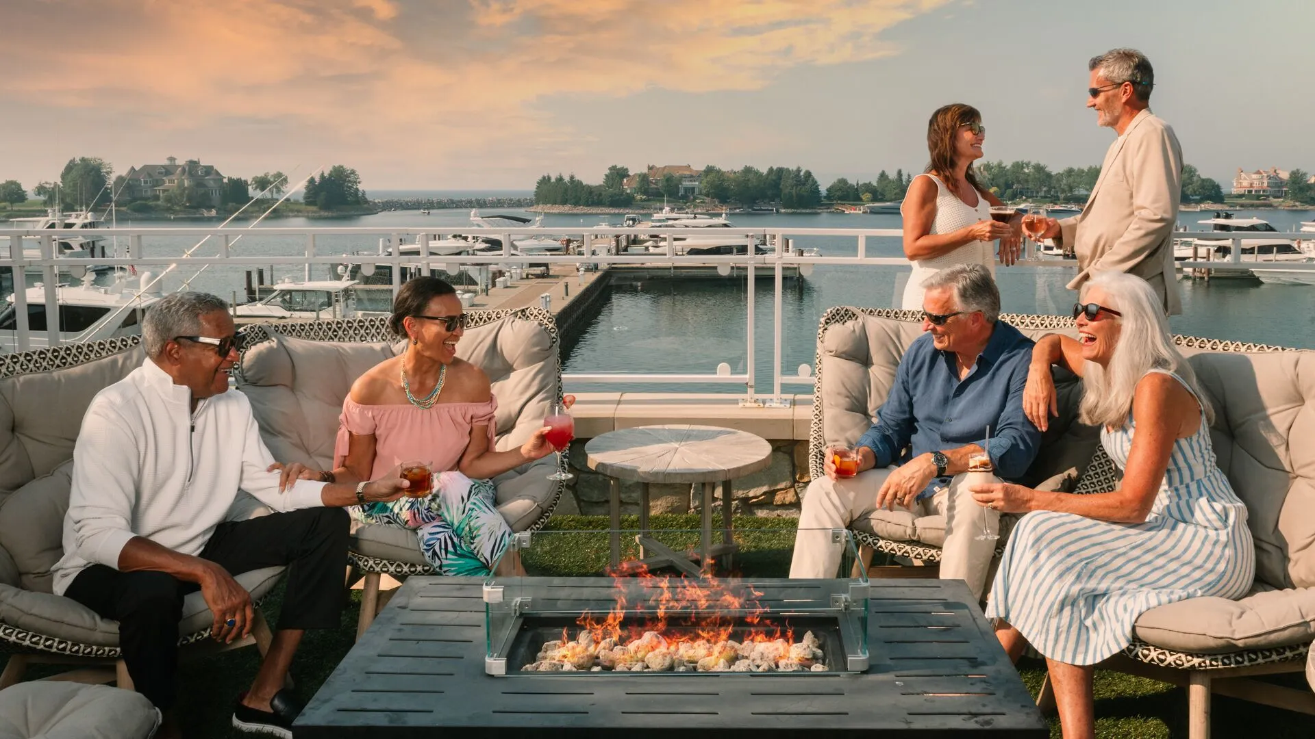This luxurious outdoor lounge area features a modern fire pit table surrounded by plush seating, perfect for hosting and socializing. The space overlooks a scenic marina filled with boats, offering stunning waterfront views during sunset. This sophisticated terrace creates an inviting atmosphere, ideal for high-end residential living.