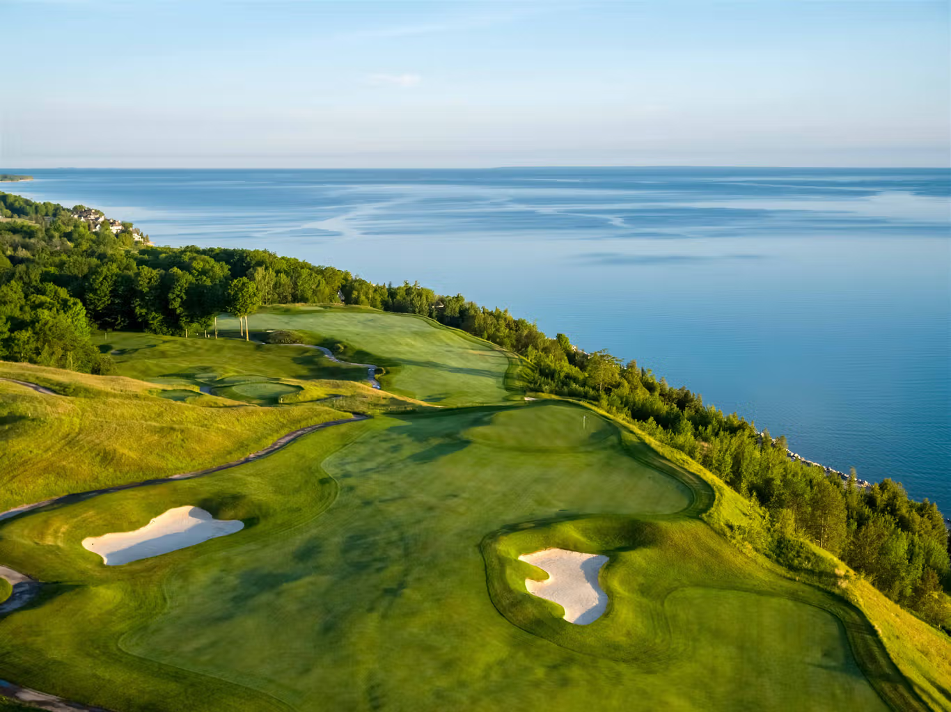 This stunning aerial view captures a premier golf course positioned dramatically along a rugged coastline. The lush green fairways and well-manicured sand bunkers contrast beautifully with the deep blue expanse of the adjacent water. The setting offers a serene, luxurious atmosphere that highlights the natural beauty and prime recreational appeal of the community.