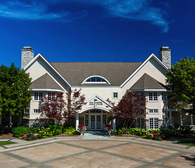 This image showcases the impressive front facade of the Bay Harbor Golf Club, featuring a grand, symmetrical architectural design with traditional gables and stone accents. The building is complemented by well-manicured landscaping, including vibrant flowers and lush trees, set against a backdrop of a clear blue sky. The welcoming entryway and expansive paved courtyard create an inviting, professional atmosphere suitable for a premier clubhouse.