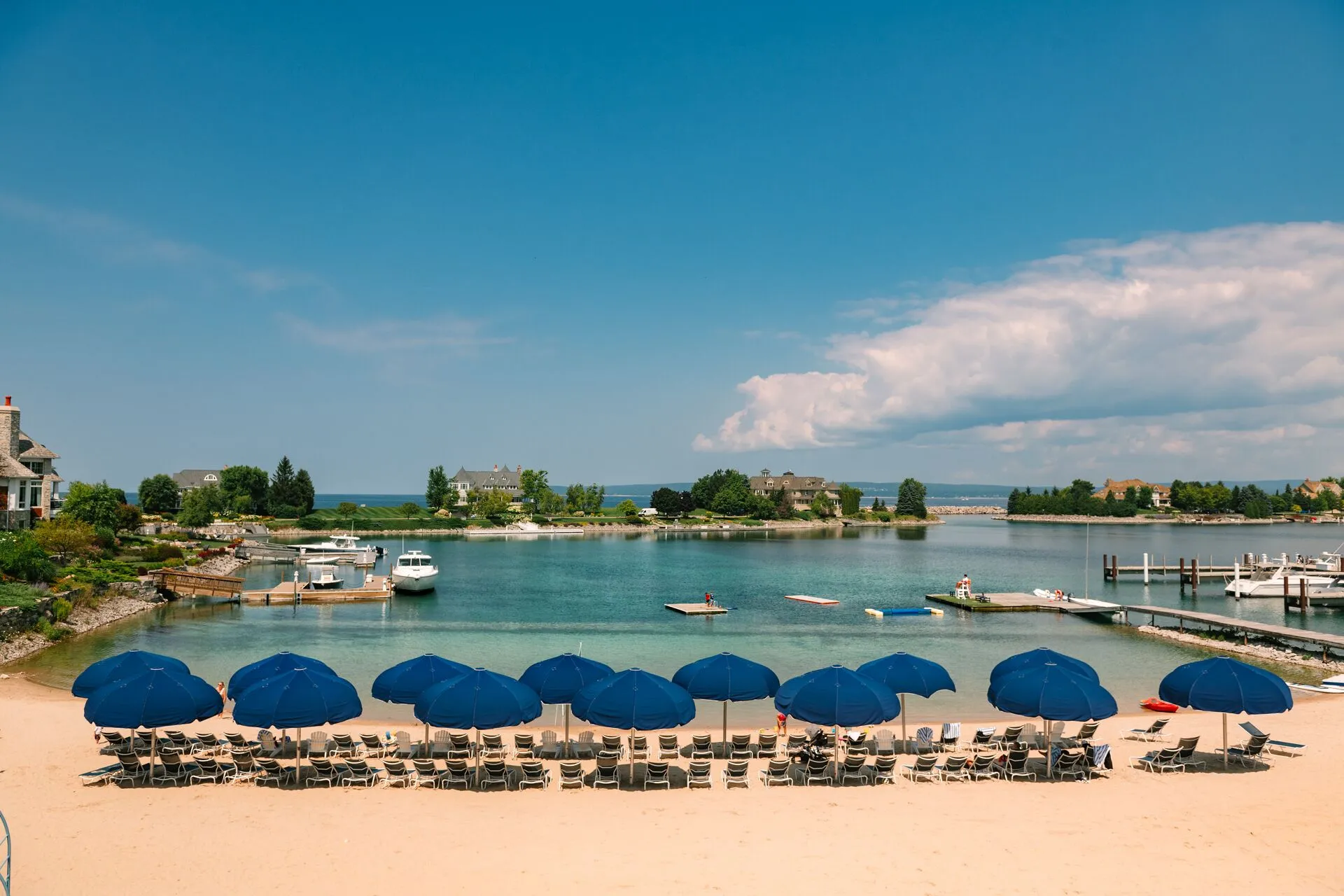 This image captures a beautifully maintained private beach area and marina, likely belonging to a luxury residential community or resort. The scene features rows of blue patio umbrellas and lounge chairs overlooking a clear, turquoise harbor with docked boats, reflecting a relaxed, upscale lifestyle. The surrounding landscape includes lush greenery, manicured lawns, and elegant waterfront homes, emphasizing the exclusivity and recreational appeal of the property.