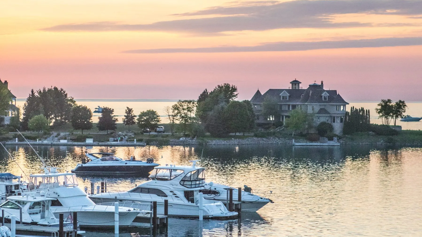 This picturesque waterfront estate offers stunning sunset views over a tranquil harbor populated by recreational yachts. The grand, traditional-style residence is situated on a landscaped point, emphasizing privacy and scenic beauty in a prime, high-end coastal or lakeside location. The setting provides a serene and luxurious atmosphere, perfect for boating enthusiasts seeking an exclusive residential retreat.