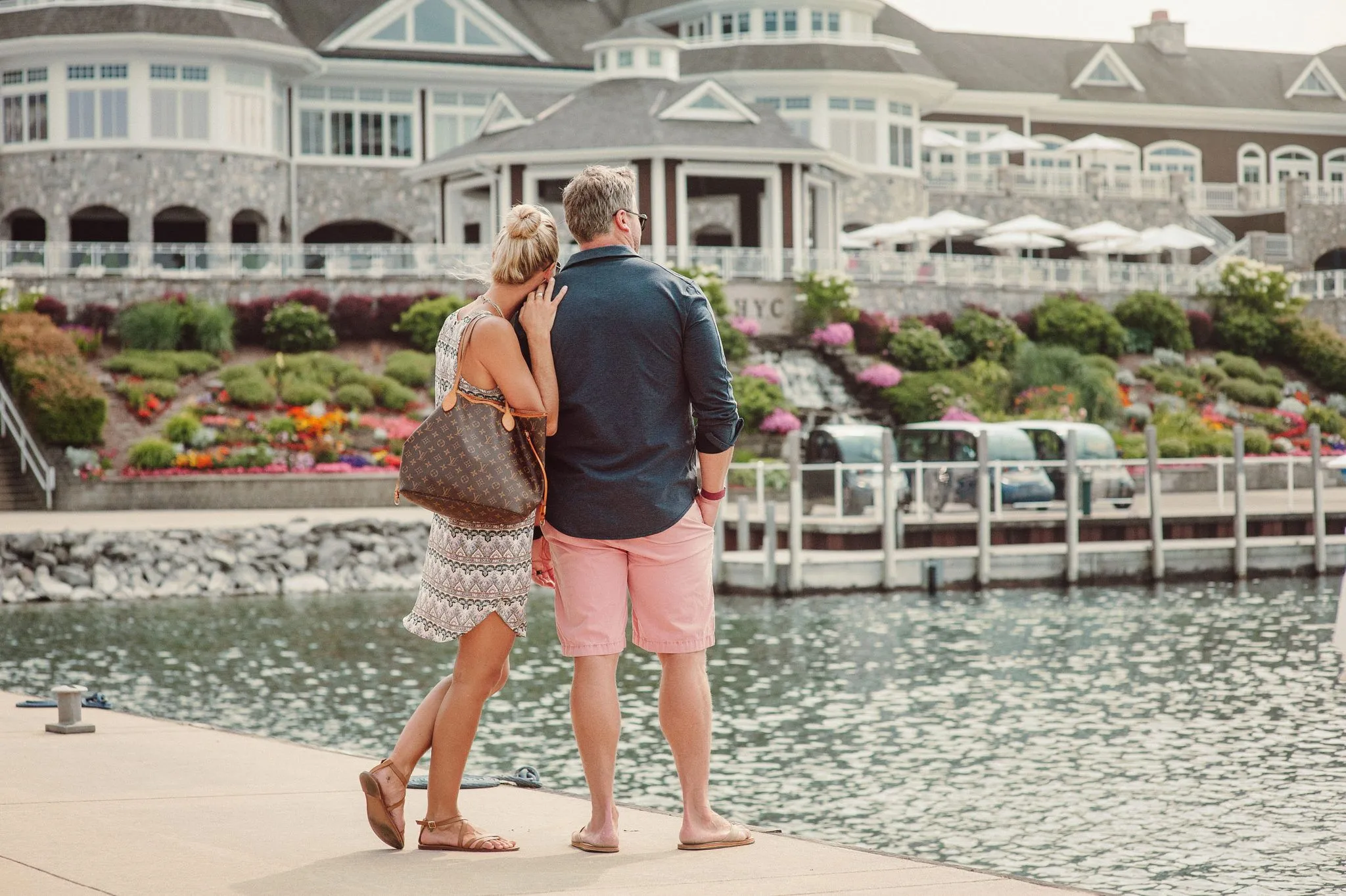 This image showcases a scenic waterfront setting featuring a large, stately clubhouse building in the background with manicured landscaping and stone accents. The foreground includes a couple standing on a concrete pier overlooking the calm water, emphasizing the relaxing, leisure-oriented lifestyle offered by this community amenity.