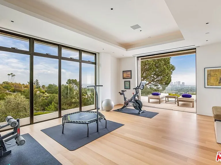 This is a bright and spacious home gym. It features large windows with beautiful views and an open layout with workout equipment. The room has light wood flooring and a clean, modern aesthetic.