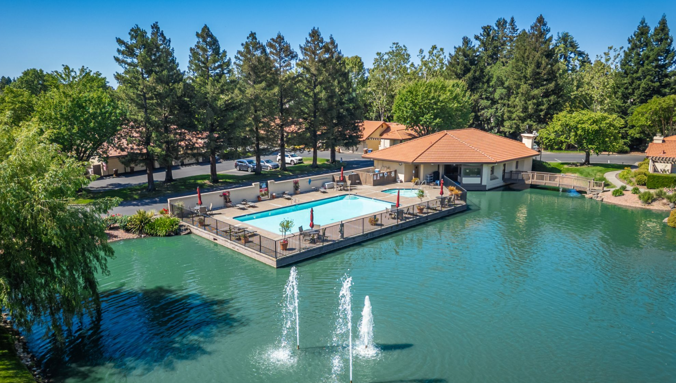 This stunning community amenity area features a swimming pool and hot tub set on a unique peninsula extending into a tranquil pond with decorative fountains. A tile-roof clubhouse provides a central gathering space, framed by lush trees and convenient paved walkways, creating a peaceful and resort-like atmosphere for residents.