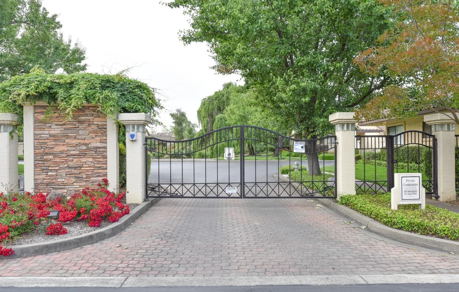 This image features the entrance to a private gated community, defined by an imposing black wrought iron gate flanked by sturdy, stone-veneer pillars. The entry is paved with decorative bricks and framed by well-maintained landscaping featuring vibrant red flowers and lush green trees, creating an inviting and secure first impression for residents and visitors alike.
