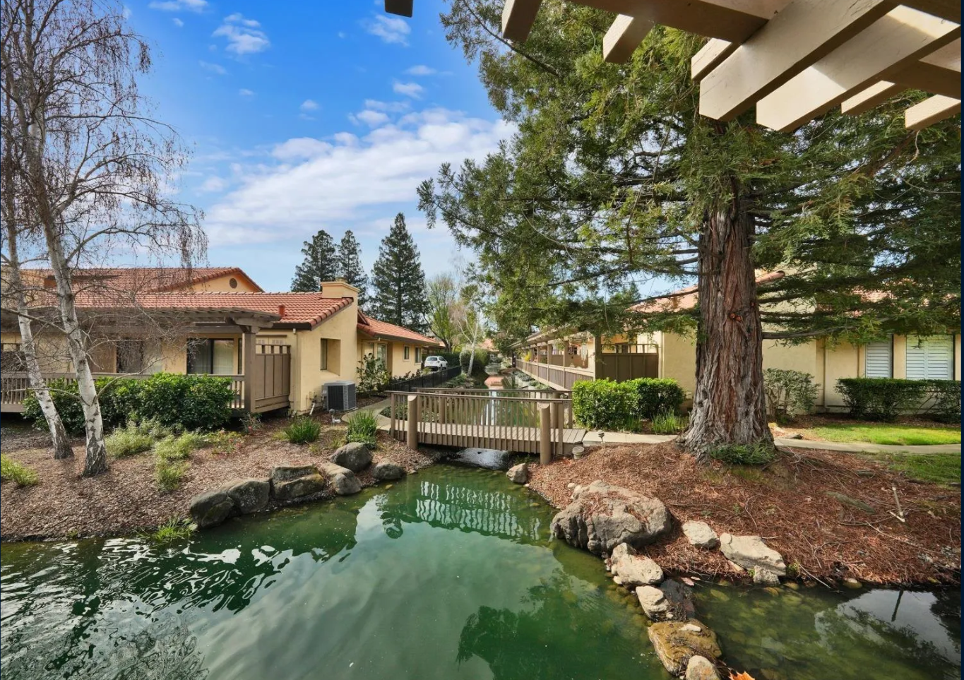 This peaceful complex setting features a serene pond crossed by a charming pedestrian footbridge, surrounded by well-maintained landscaping and mature trees. The architecture showcases a cohesive Mediterranean-inspired design with stucco siding and terracotta tile roofs. The tranquil water feature and lush green spaces provide a picturesque and relaxing atmosphere for residents.