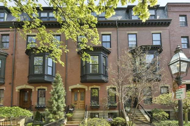 This image showcases the front exterior of a row of townhouses. The buildings feature classic brick facades and bay windows, exuding traditional charm. The entryway of one of the houses is visible, leading up to a wooden door with brass accents, and a vintage-style lamppost stands nearby, enhancing the neighborhood's appeal.