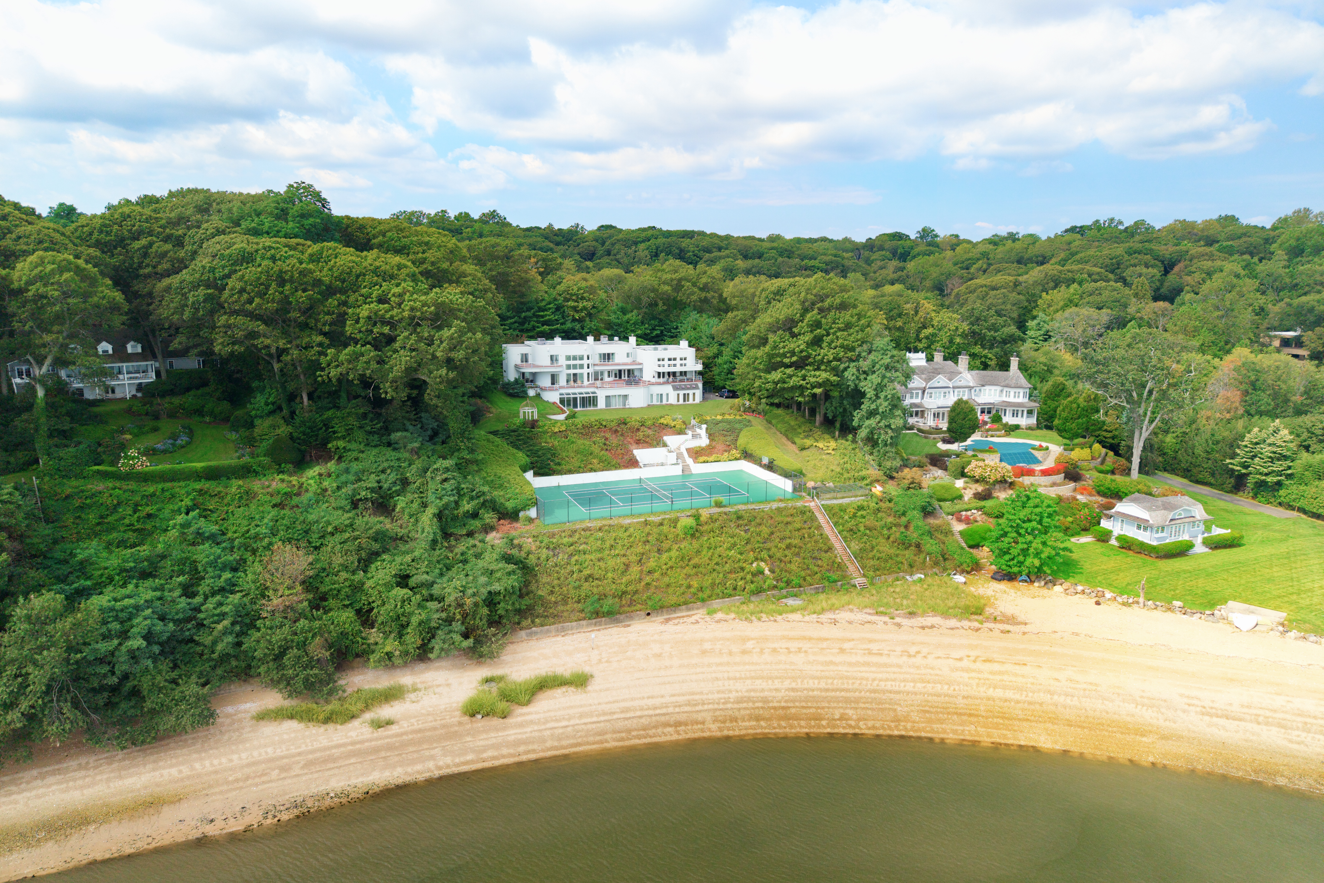 This high-angle aerial view captures a sprawling coastal estate nestled within a lush, wooded landscape overlooking a sandy beach and calm water. The property features a prominent modern white residence, a private tennis court, and a secondary traditional home with a swimming pool, all integrated into the natural hillside. The perspective emphasizes the expansive, secluded nature of the grounds and the seamless transition from the manicured lawns to the shoreline.