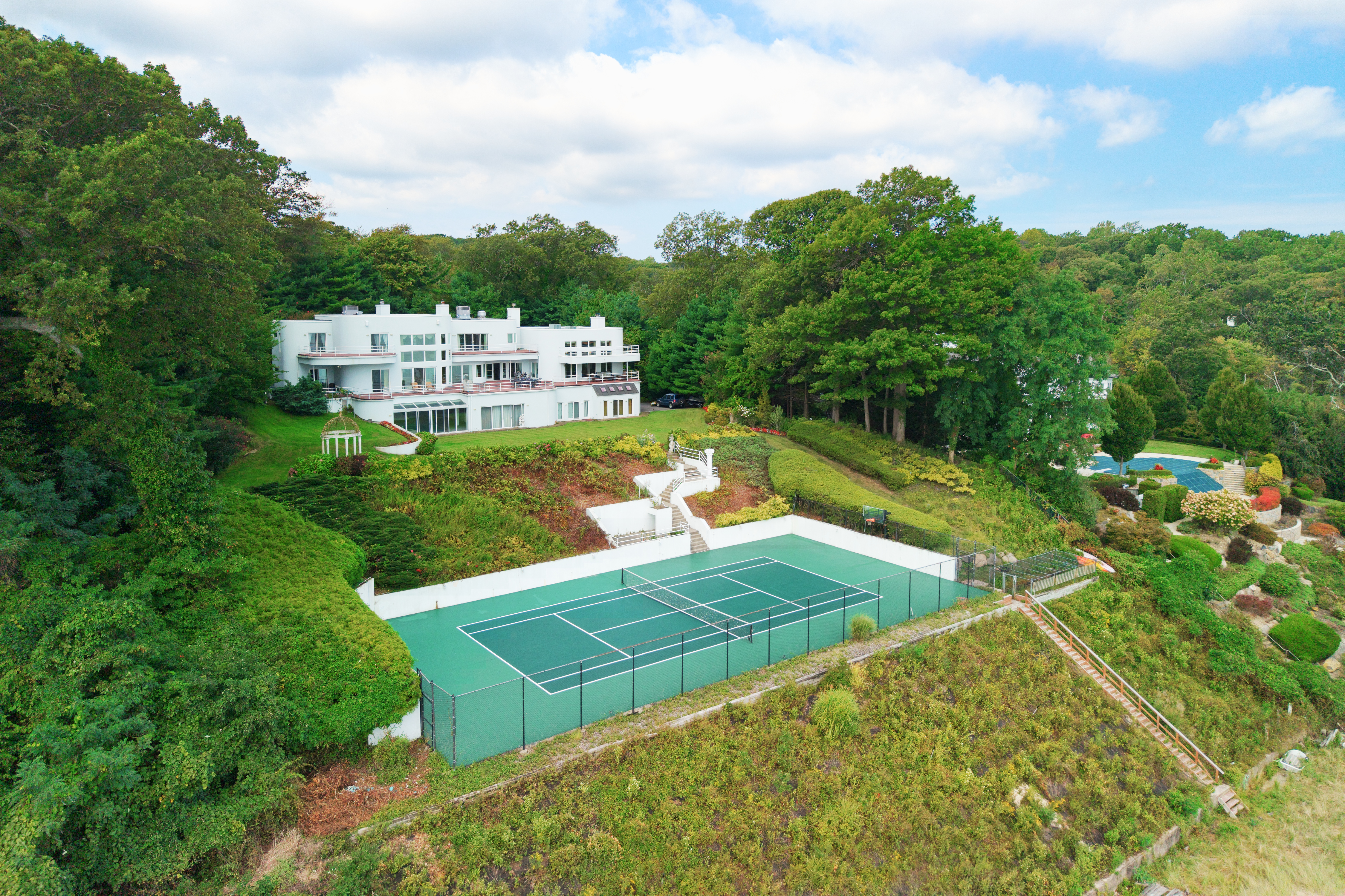 This high-angle aerial shot captures a sprawling, modern white estate nestled into a lush, wooded hillside. The property features a prominent tennis court in the foreground, tiered landscaping with stone stairs, and a large swimming pool visible in the distance. The architectural style is sleek and contemporary, emphasizing expansive outdoor living spaces and integration with the natural environment.