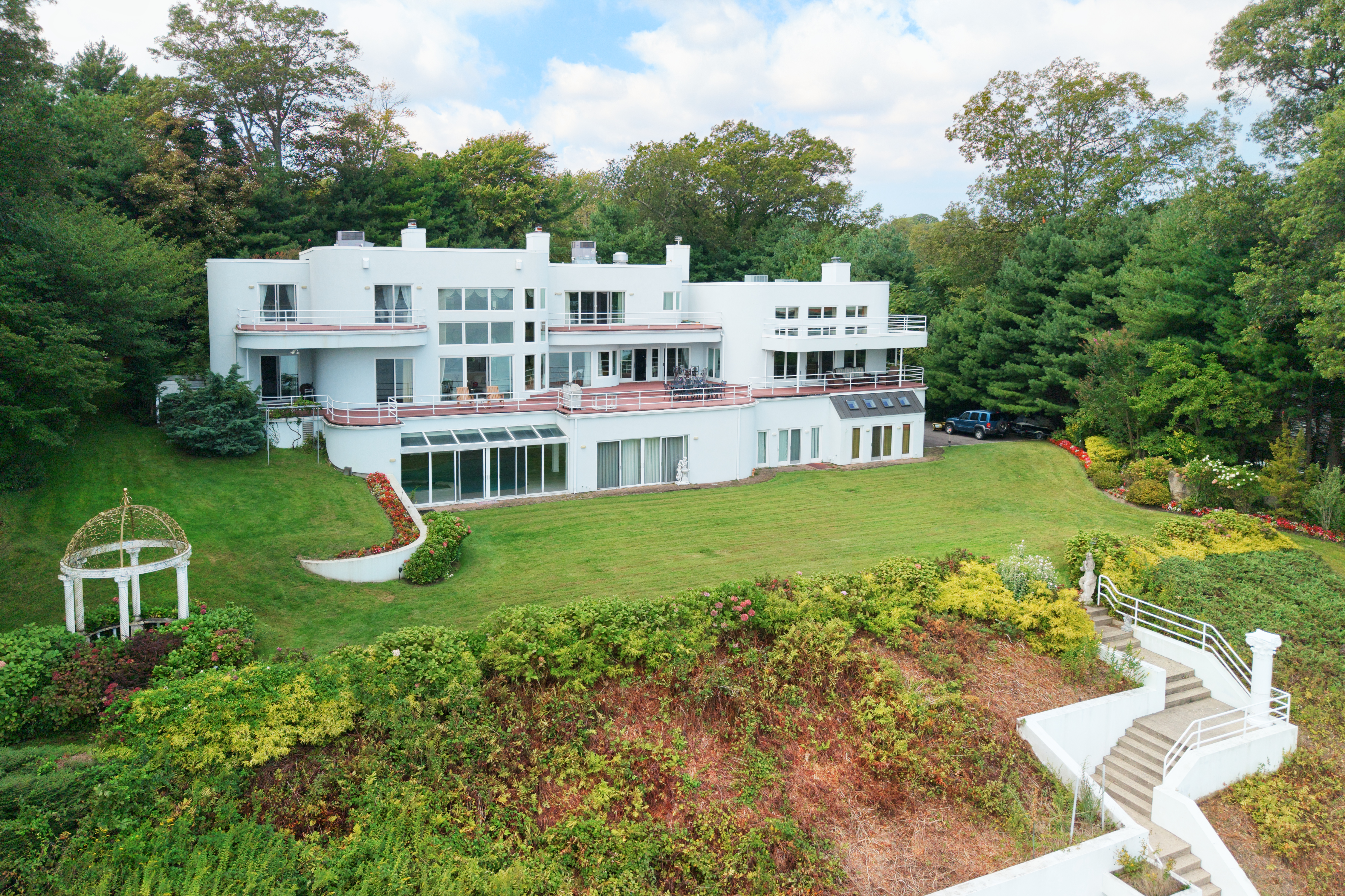 This expansive, multi-level contemporary estate features a striking white facade with curved architectural lines and multiple balconies overlooking a lush, manicured lawn. The property is nestled against a dense forest backdrop, offering a private and serene atmosphere with a decorative gazebo and stone-stepped pathways. The elevated perspective captures the grand scale of the residence, emphasizing its seamless integration with the surrounding natural landscape.