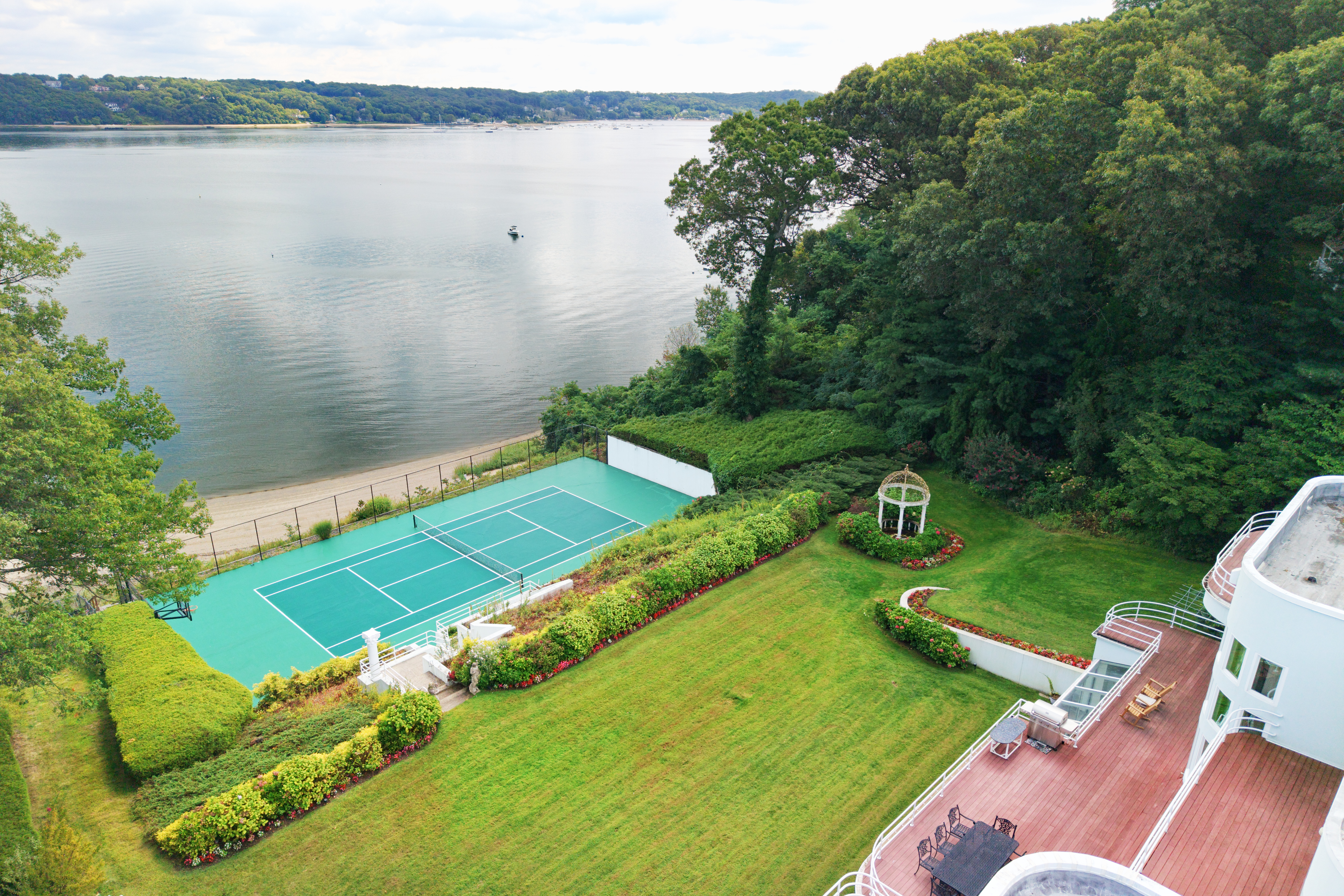 This stunning aerial perspective captures a luxurious waterfront estate featuring a private tennis court nestled between a lush green lawn and a sandy beach. The property includes a spacious wooden deck with outdoor dining and grilling areas, seamlessly blending indoor-outdoor living with panoramic views of the bay. The scene is framed by mature trees and manicured landscaping, conveying an atmosphere of exclusive coastal elegance.