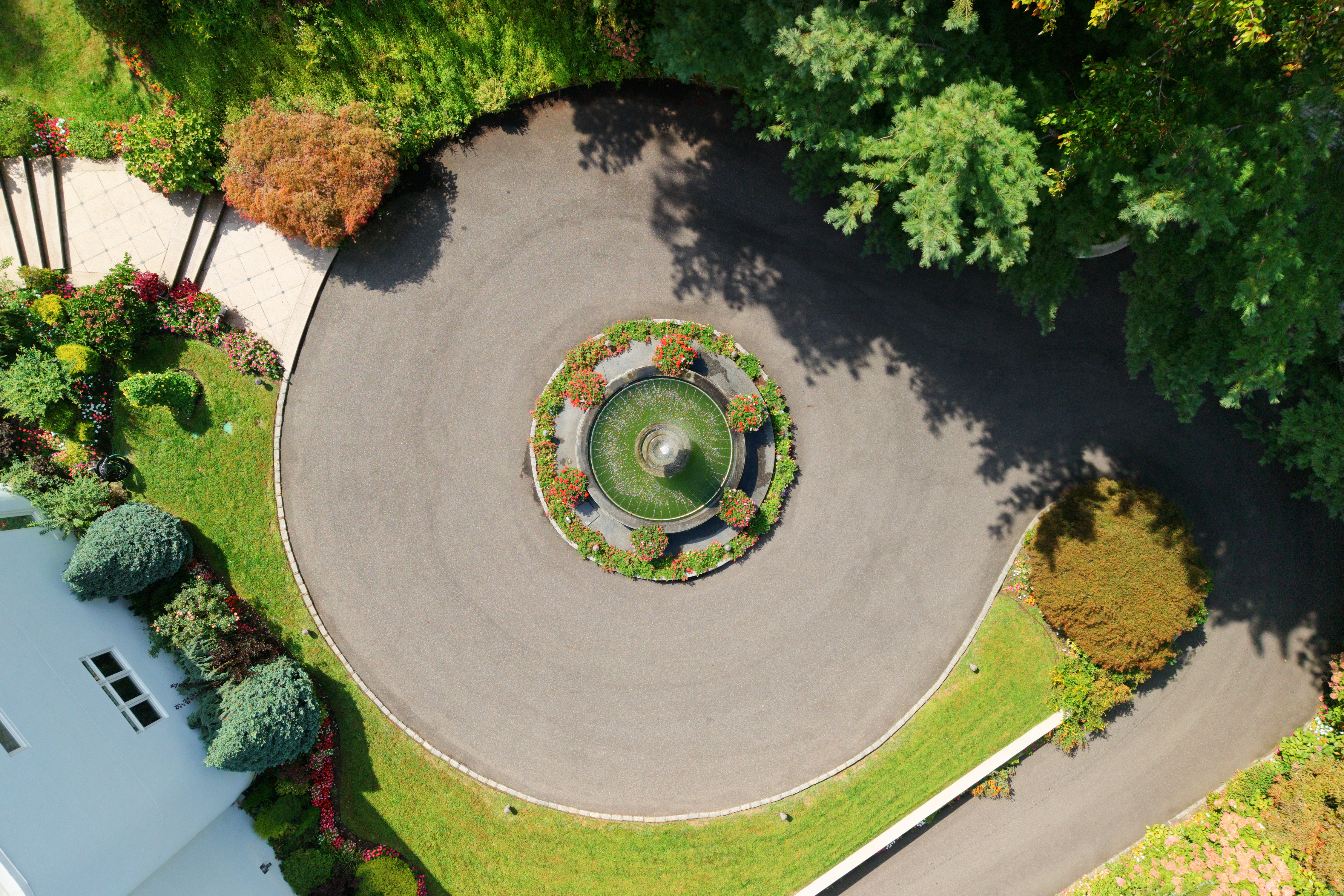 This high-angle aerial shot captures a beautifully landscaped circular driveway centered around a classic stone fountain adorned with vibrant red flowers. The property features lush green lawns, mature trees, and manicured garden beds, creating a serene and elegant arrival experience. The perspective emphasizes the symmetrical layout and the well-maintained grounds of the estate.