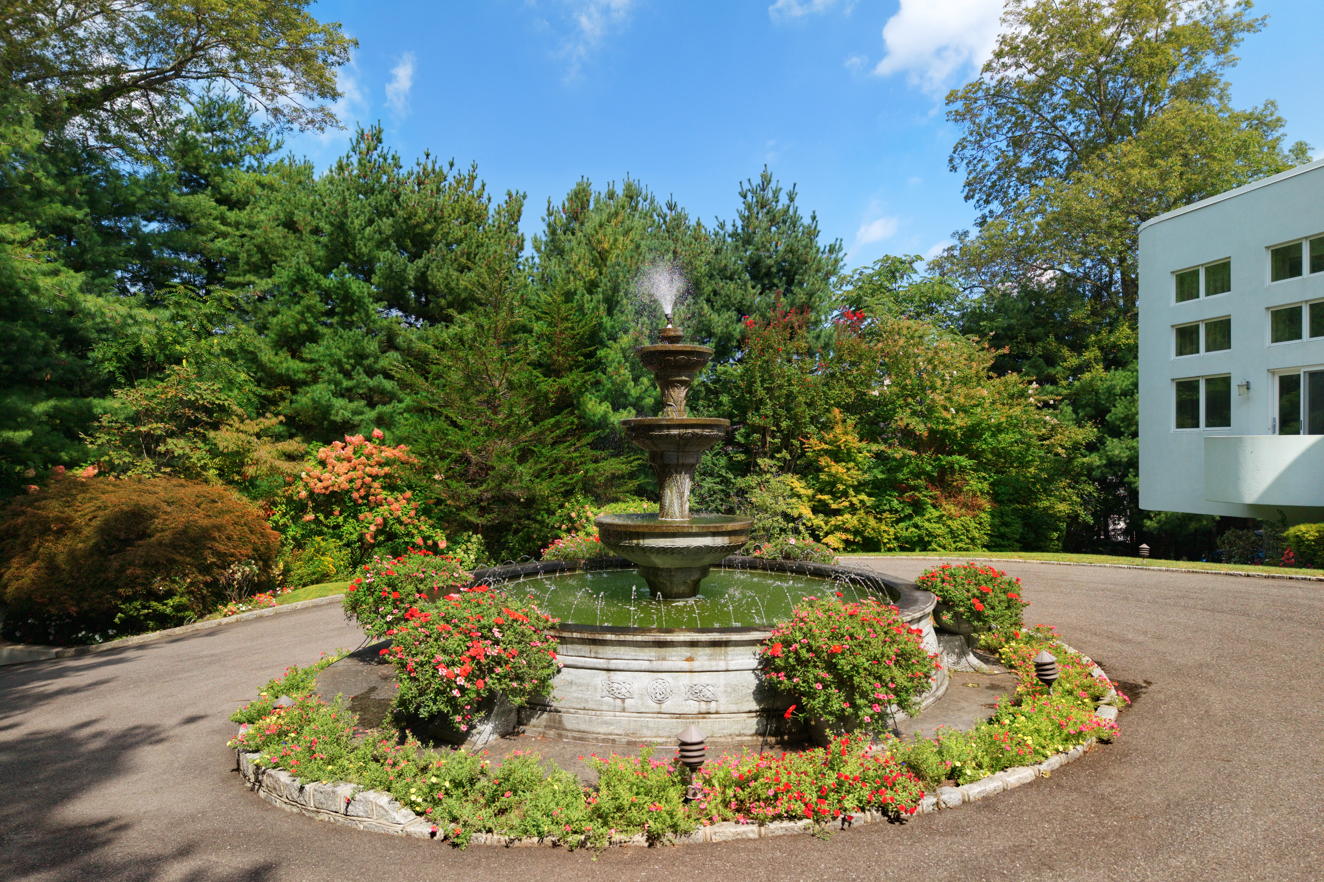 This image captures a beautifully landscaped circular driveway featuring a classic three-tiered stone fountain as its centerpiece. The fountain is surrounded by vibrant red flowers and lush greenery, creating a serene and elegant focal point. The perspective is a ground-level shot that emphasizes the manicured garden setting and the inviting, upscale atmosphere of the property's exterior.