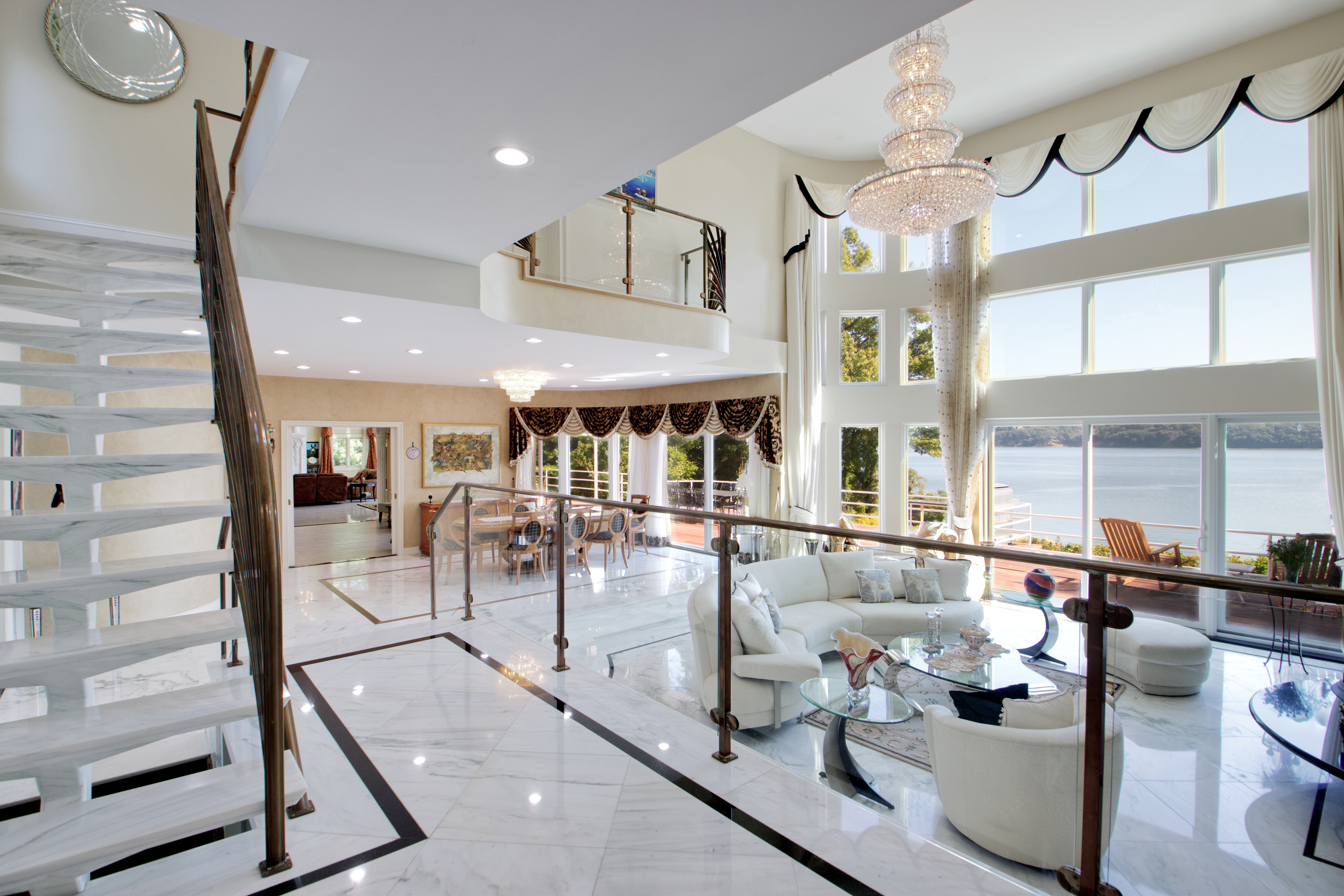 This expansive, high-ceilinged living area features a luxurious open-concept design with polished marble flooring and a grand crystal chandelier. The space is anchored by a curved white sofa set overlooking a stunning waterfront view through floor-to-ceiling windows, while a floating staircase adds a dramatic architectural element. The overall impression is one of sophisticated, high-end elegance with a seamless transition between indoor living and the natural scenery outside.
