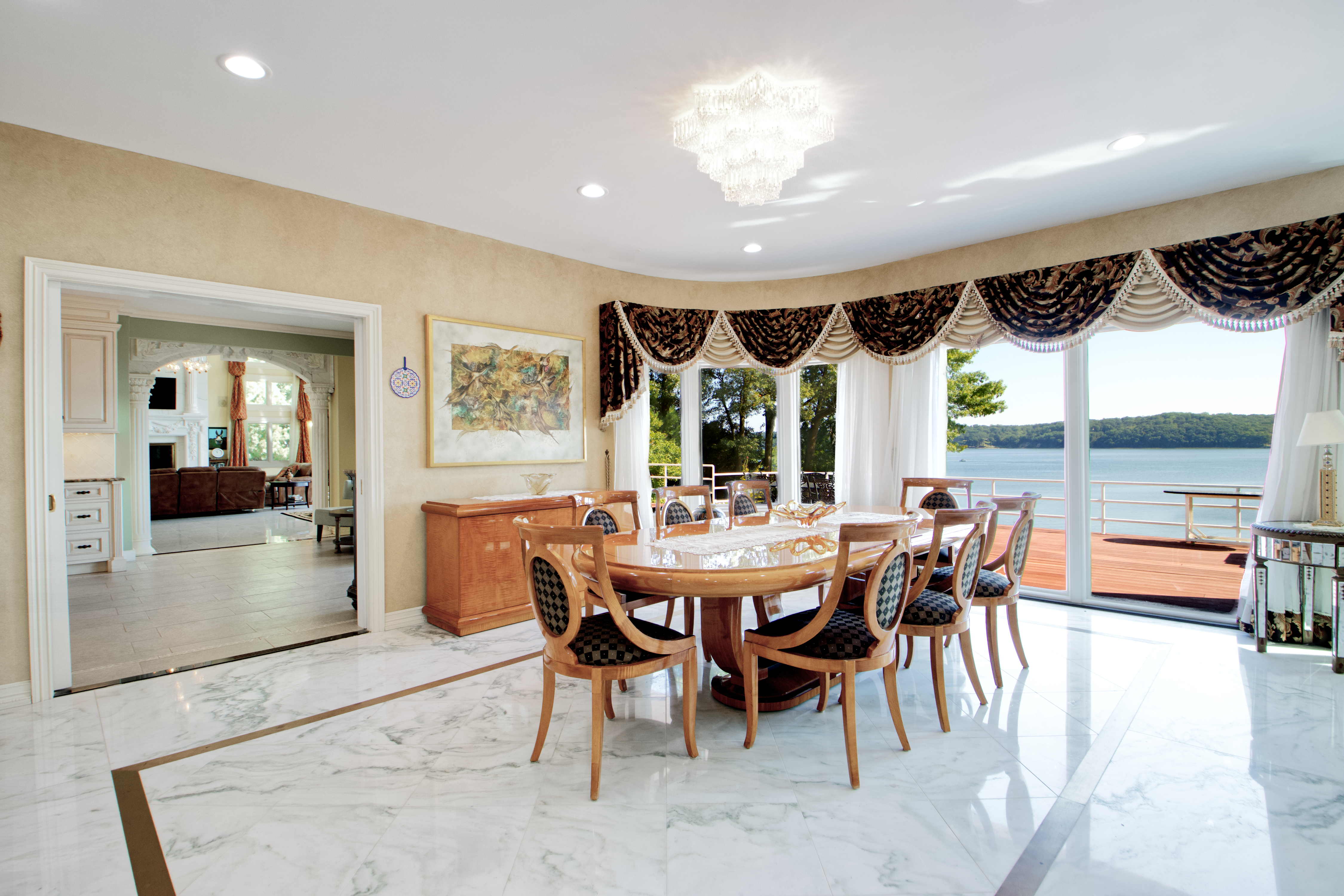 This elegant dining room features a polished marble floor with a dark inlay border and a large, oval wooden dining table surrounded by eight upholstered chairs. A crystal chandelier hangs above the table, while a curved wall of windows offers a stunning, unobstructed view of the lake and deck. The room is decorated in a traditional, formal style with ornate window valances and a view into an adjacent living area with a fireplace.