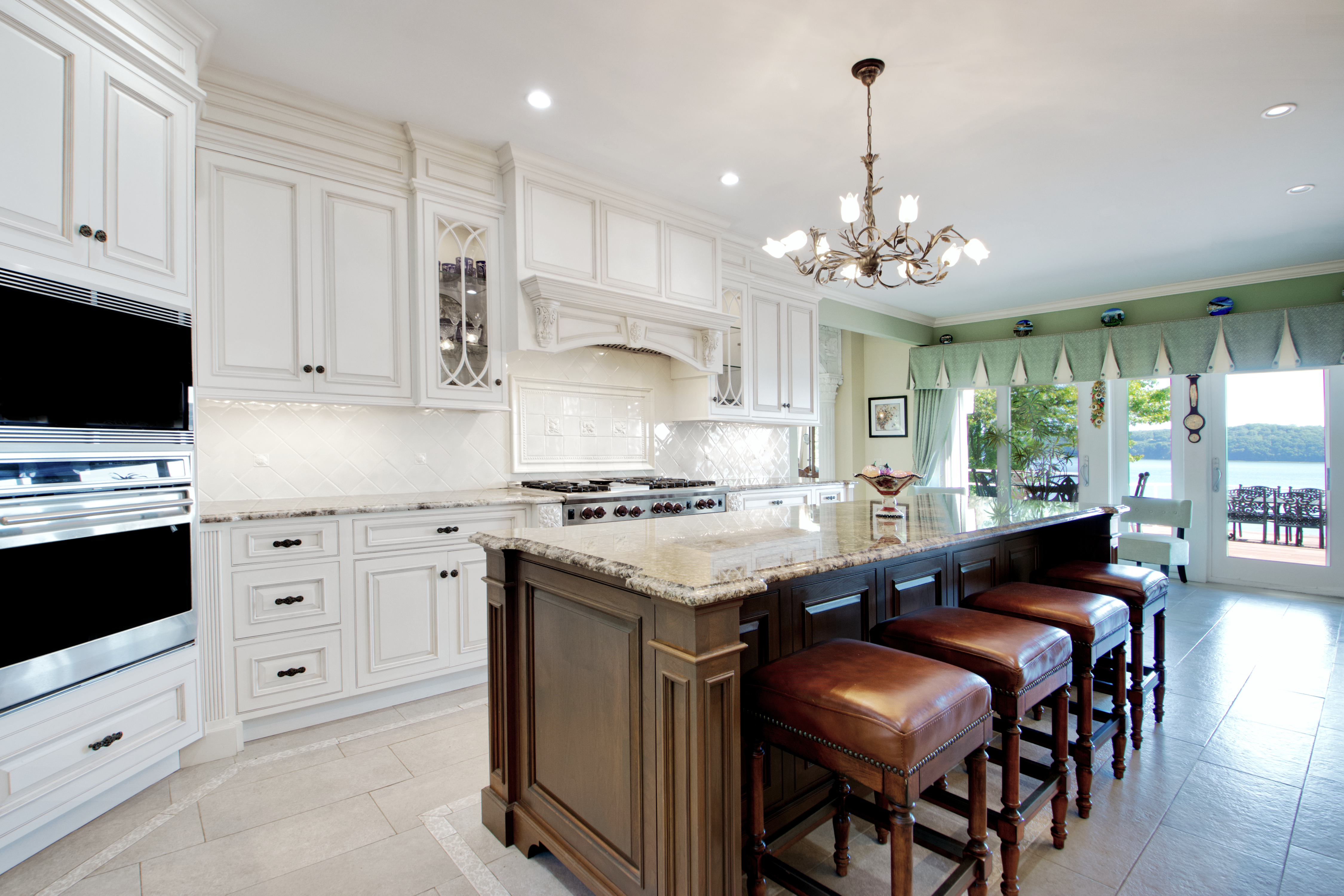 This elegant, high-end kitchen features a sophisticated contrast between white perimeter cabinetry and a dark wood center island topped with granite. The space is illuminated by a decorative chandelier and recessed lighting, while large glass doors provide a seamless transition to an outdoor patio with a scenic water view. The overall aesthetic is traditional and luxurious, emphasizing high-quality finishes and a spacious, open-concept layout.