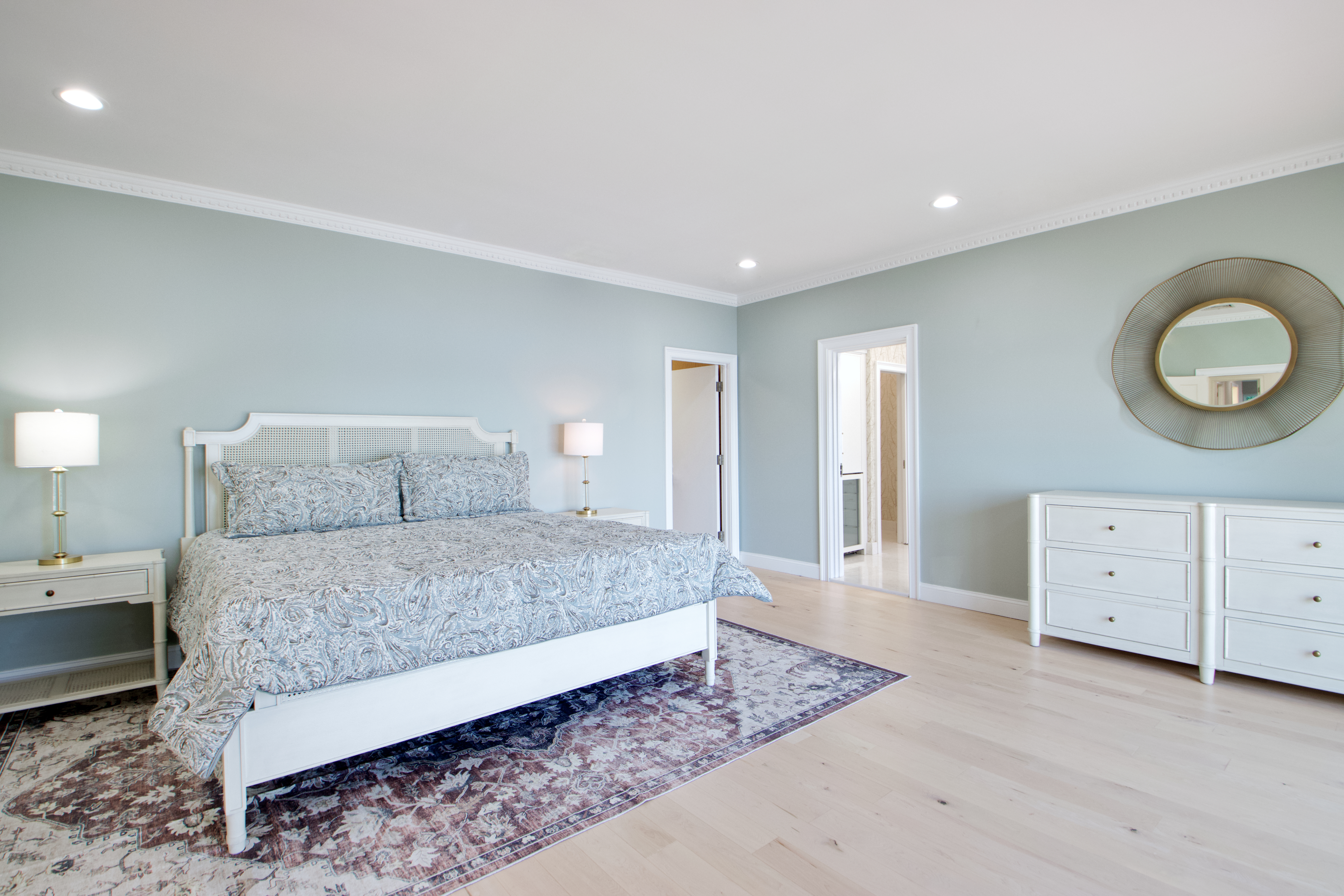 This spacious primary bedroom features light blue walls, light hardwood flooring, and a large bed with a white frame and patterned bedding. A circular gold-rimmed mirror hangs above a white dresser, while two matching nightstands with lamps flank the bed. The room is bright and airy, with a wide-angle perspective that highlights the clean, transitional design and the transition into an adjacent hallway.